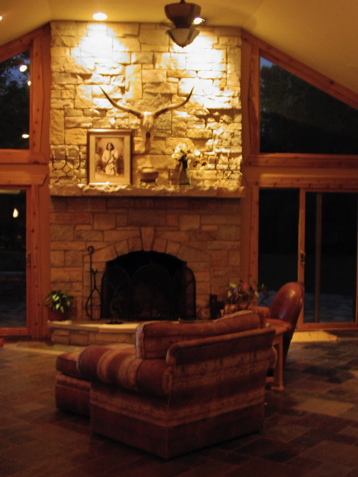 Living room with a stone fireplace, a mounted deer skull with antlers above the fireplace, a framed picture, a bouquet of flowers, and leather furniture near large windows.