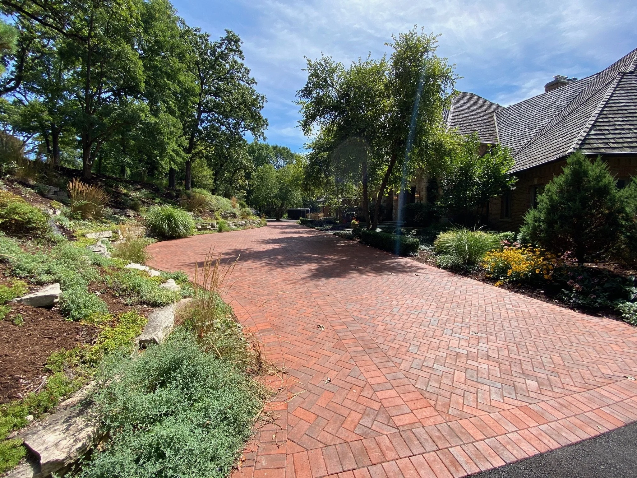 A brick driveway in front of a house with a steeply sloped roof, surrounded by trees and bushes on a sunny day with a blue sky.