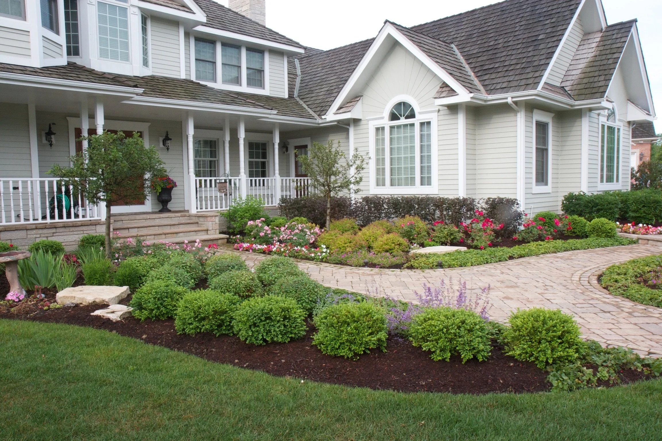 A large two-story house with white siding and a shingled roof, surrounded by a landscaped garden with colorful flowers, green shrubs, and a brick pathway.