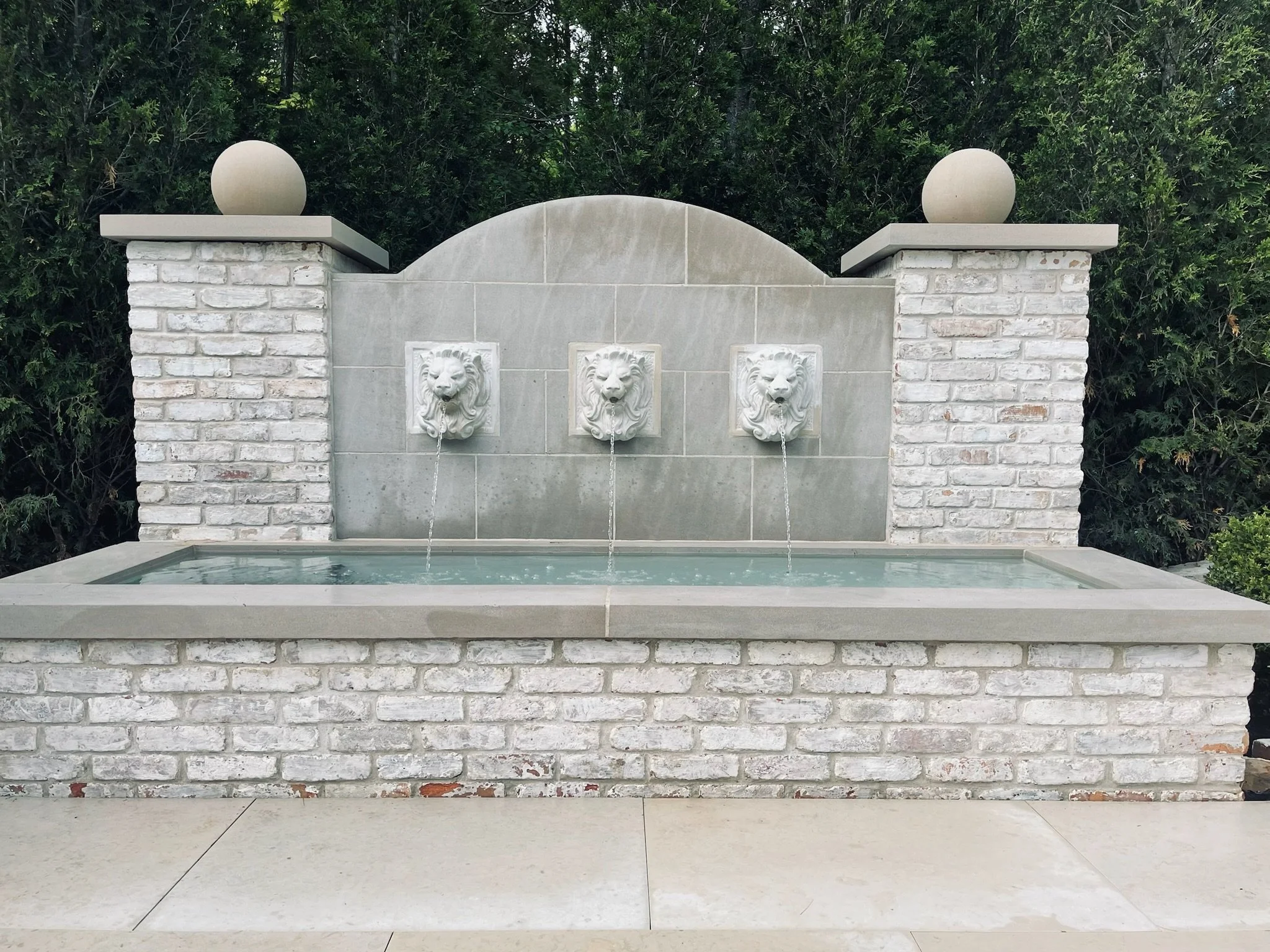 A stone wall fountain with three lion head water spouts, white bricks, and a concrete top with two spherical finials, surrounded by green shrubbery.