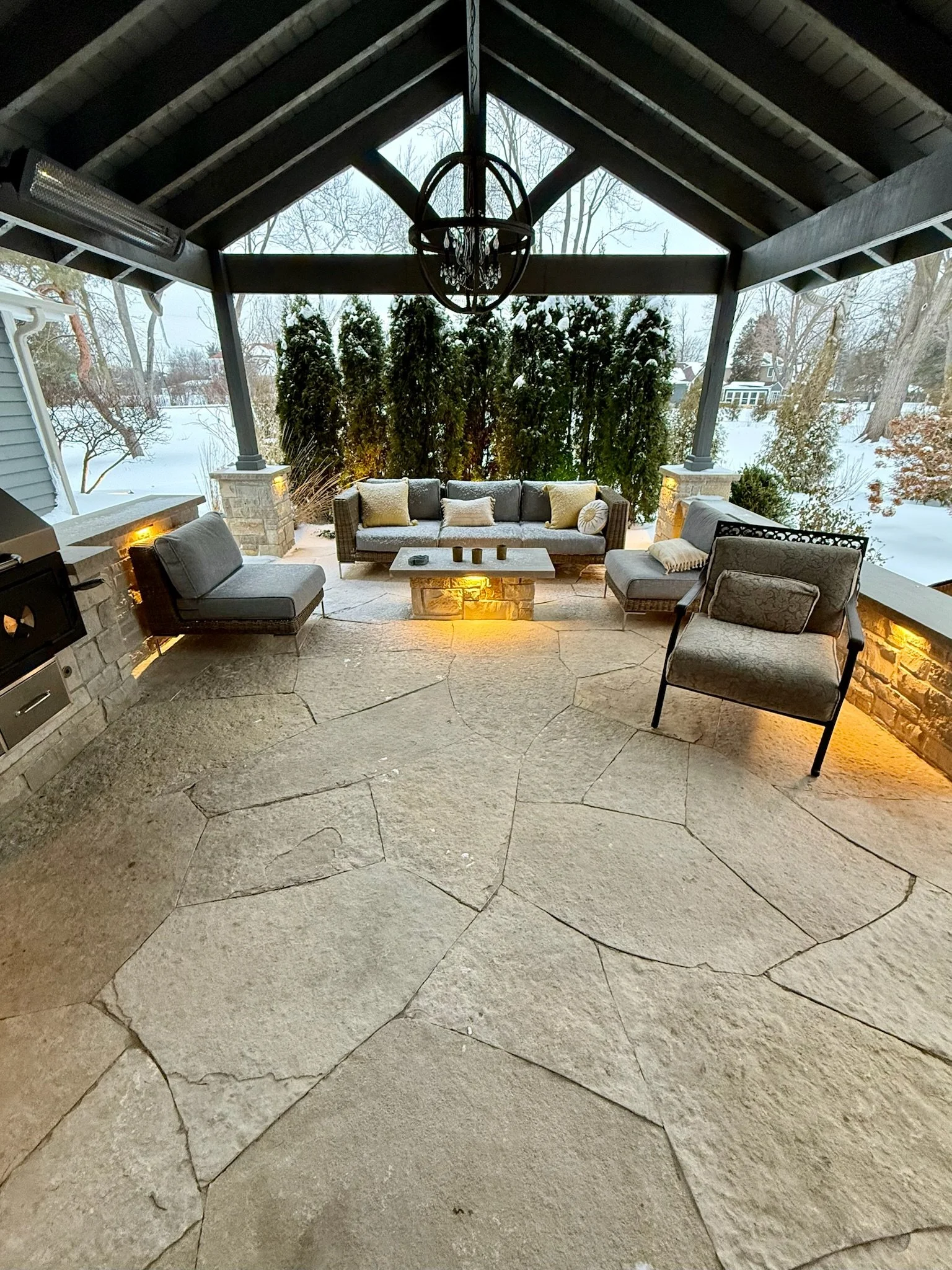 Outdoor patio with stone flooring, seating area with sofa and chairs, stone and wood built-in features, snow-covered landscape, trees, and houses in the background, illuminated with warm lighting.
