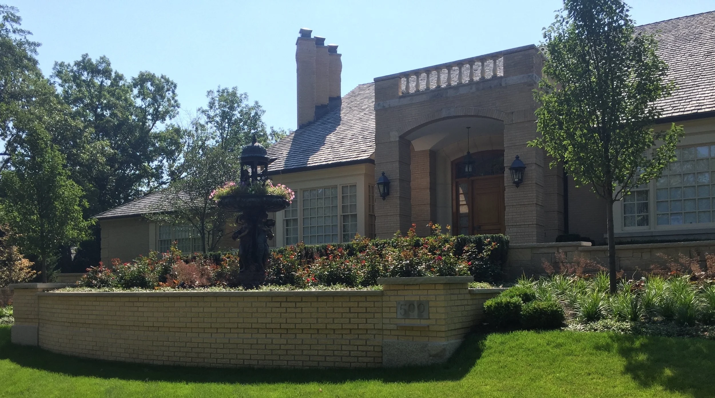 Front view of a large house with a brick exterior, multiple windows, a chimney, and a well-maintained garden with trees, bushes, and a decorative fountain with flowers.