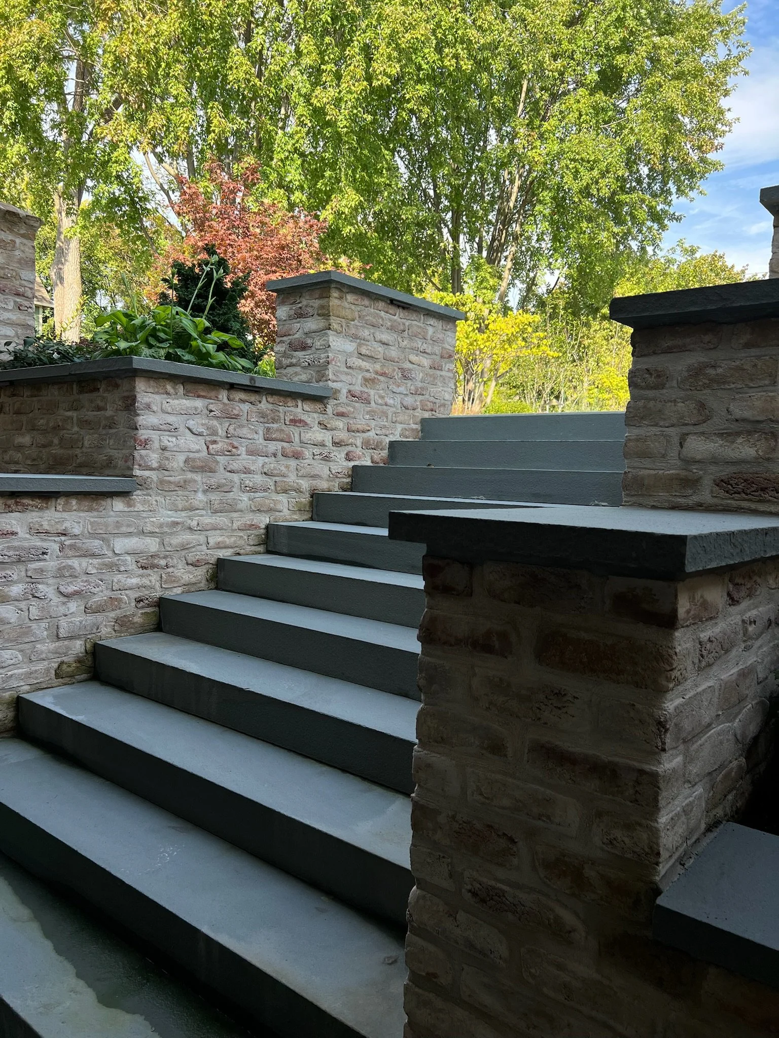 Brick steps leading up to a higher level with a brick wall and lush green trees in the background.