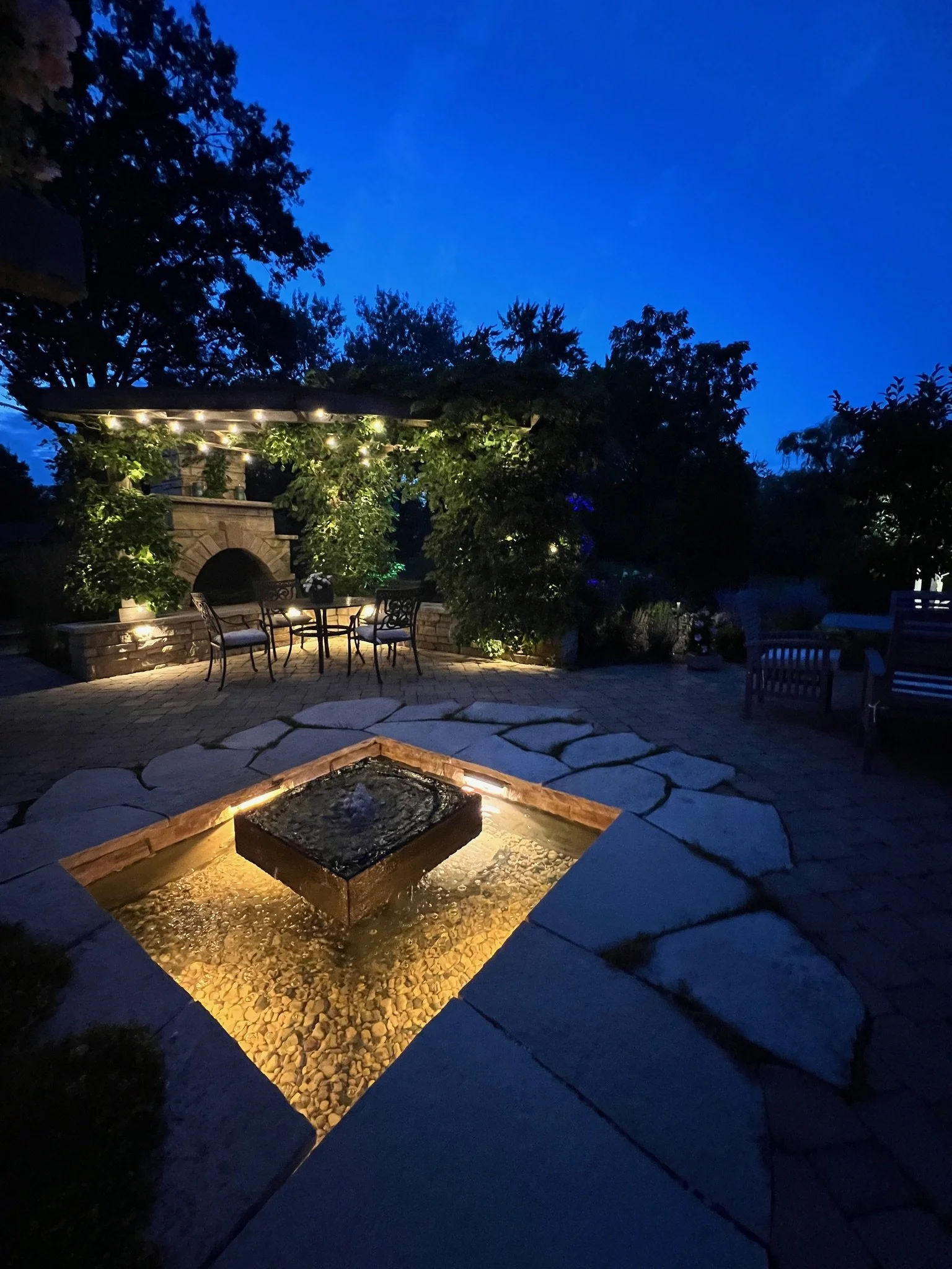An outdoor patio area at night with a stone fire pit, surrounded by chairs, a stone fireplace with string lights, trees and shrubs, and a dark blue sky in the background.