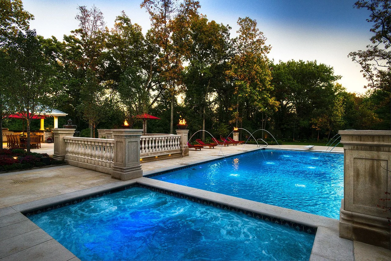 A luxurious backyard swimming pool at dusk with trees in the background, red lounge chairs, and a patio area with tables, umbrellas, and torches.