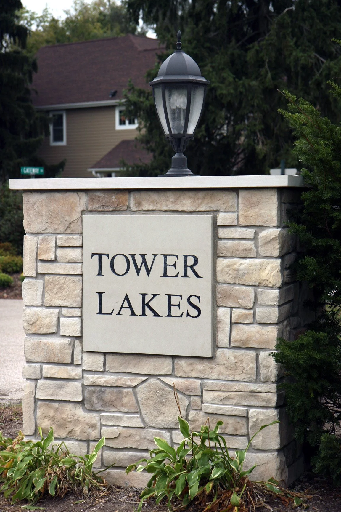 A stone entrance pillar with a sign reading 'TOWER LAKES' and a black lamp post on top, surrounded by greenery.
