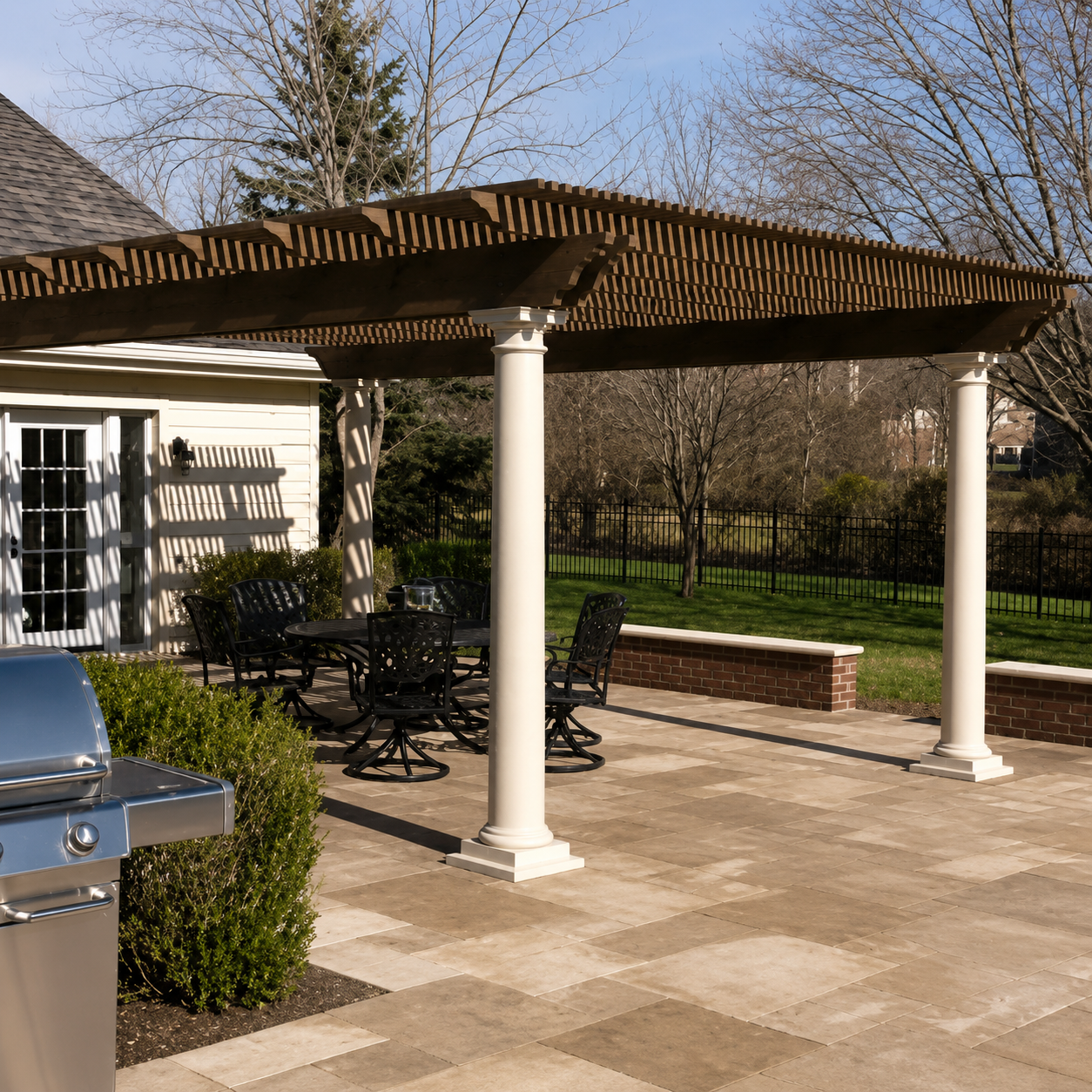 Backyard patio with a wooden pergola, outdoor dining table with chairs, a grill, and a grassy yard with trees and a black metal fence.