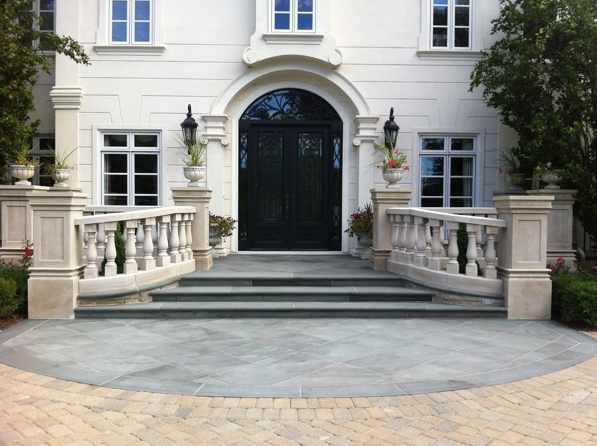 Front view of a white house with two large windows above and a black ornate gate entrance, flanked by planters with flowers and tall plants, with a stone pathway and railing in the foreground.