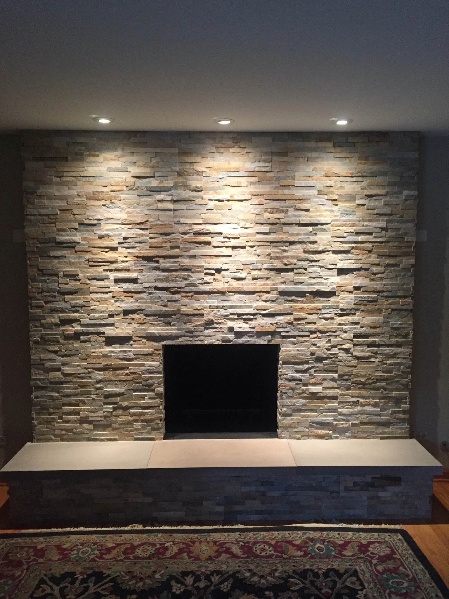 Interior of a room with a large stone fireplace, three ceiling lights above, a decorative rug, and a flat panel in the center of the fireplace.