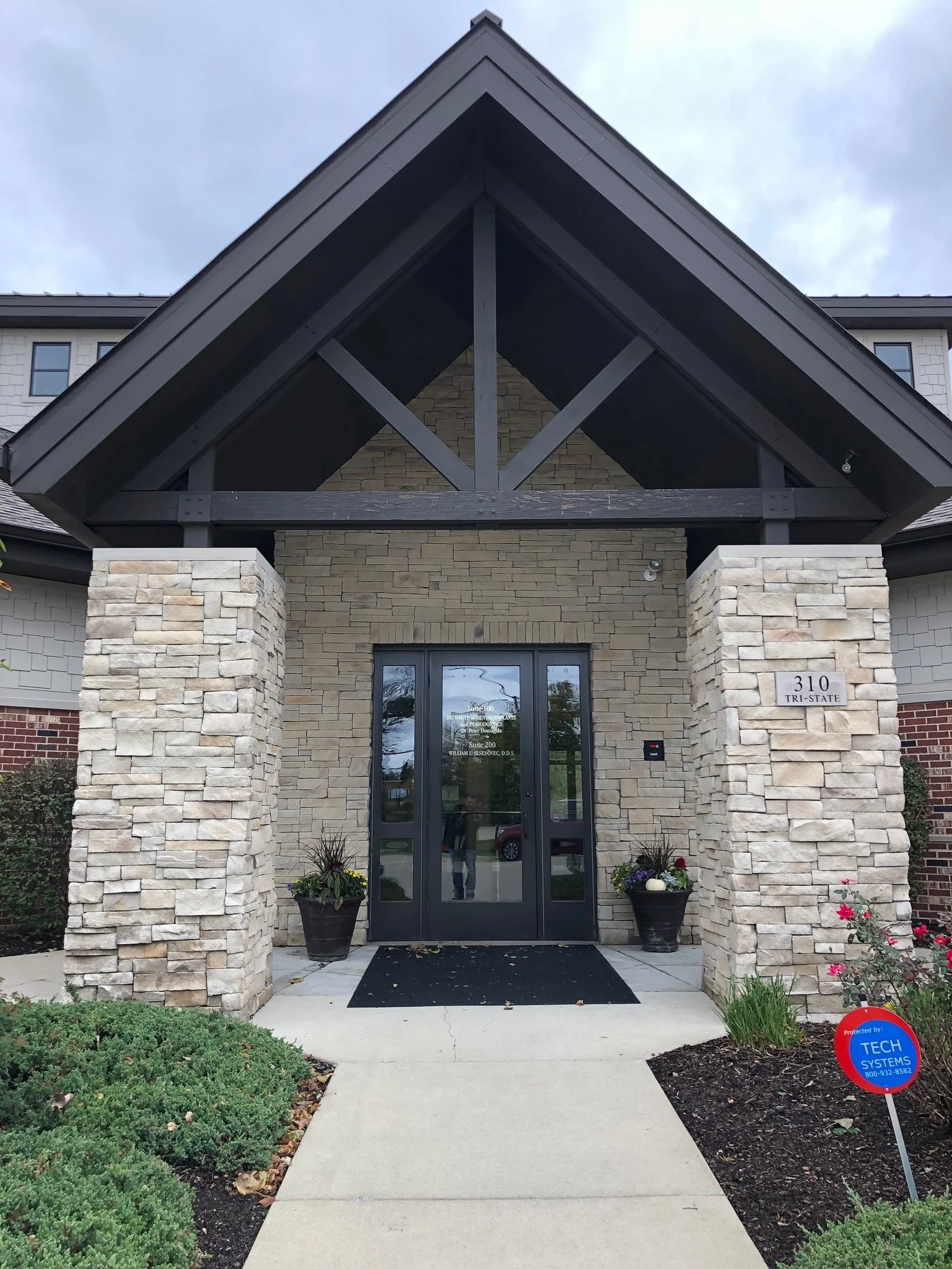 Entrance of a building with stone pillars, glass double doors, a black doormat, potted flowers, and a sign that reads '310 TRI-STATE'. There is a security camera on the right side and a blue sign with white text on the grass indicating a protected si
