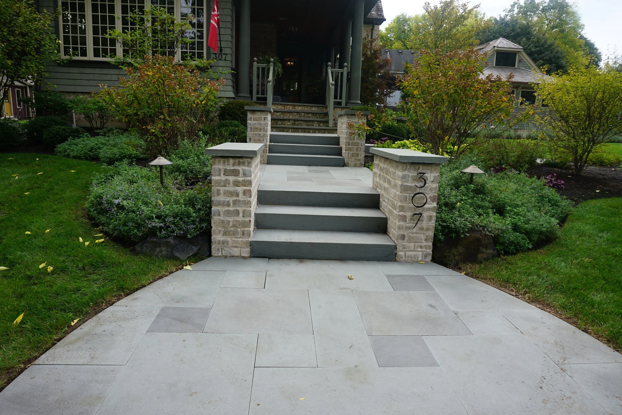 Front porch of a house with stairs, brick posts, and black railing, surrounded by green bushes and trees, with neighboring houses visible in the background.
