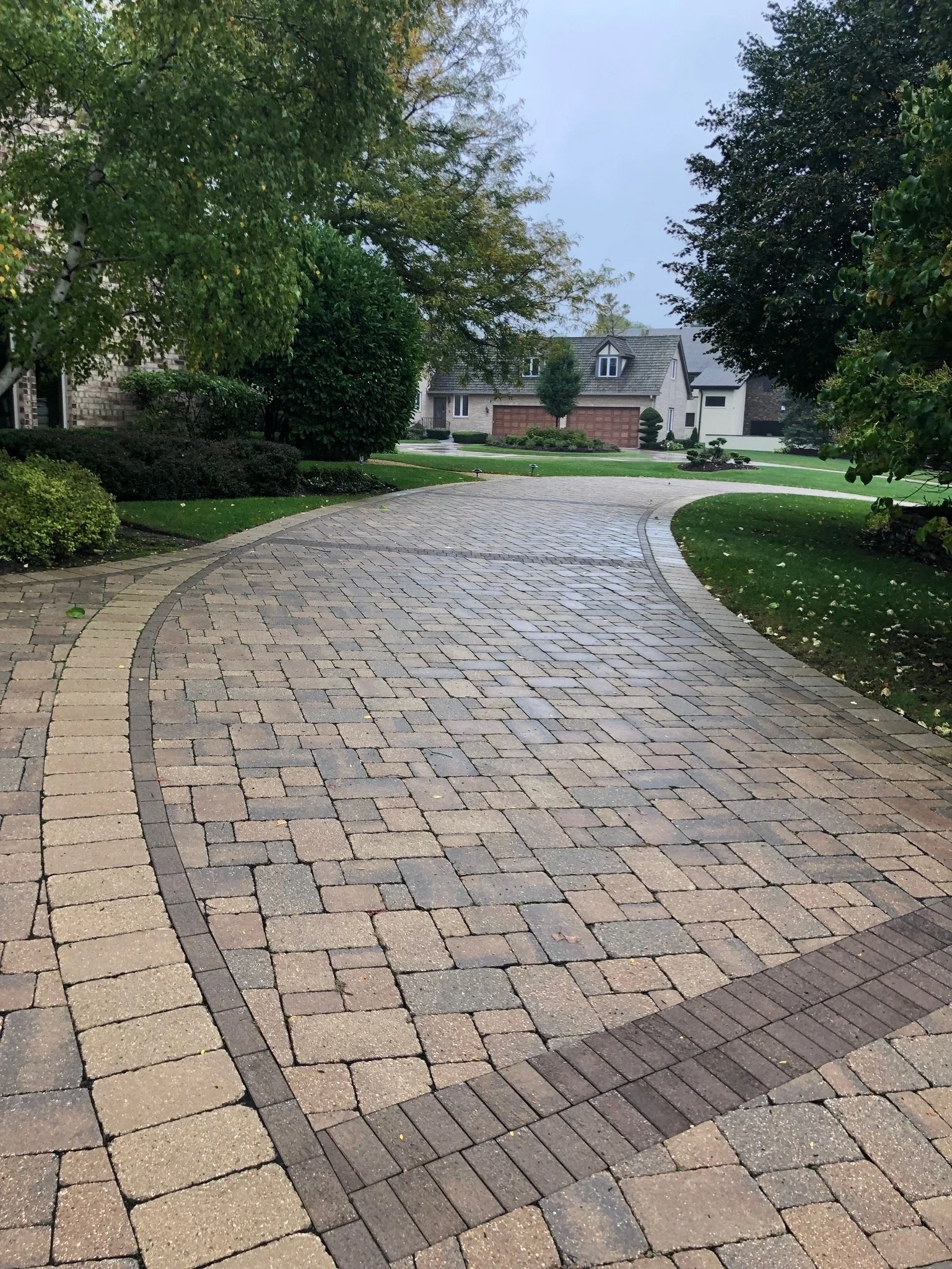 A residential neighborhood with a paved walkway, green grass, and trees. In the background, there are houses with garages, and the sky appears overcast.