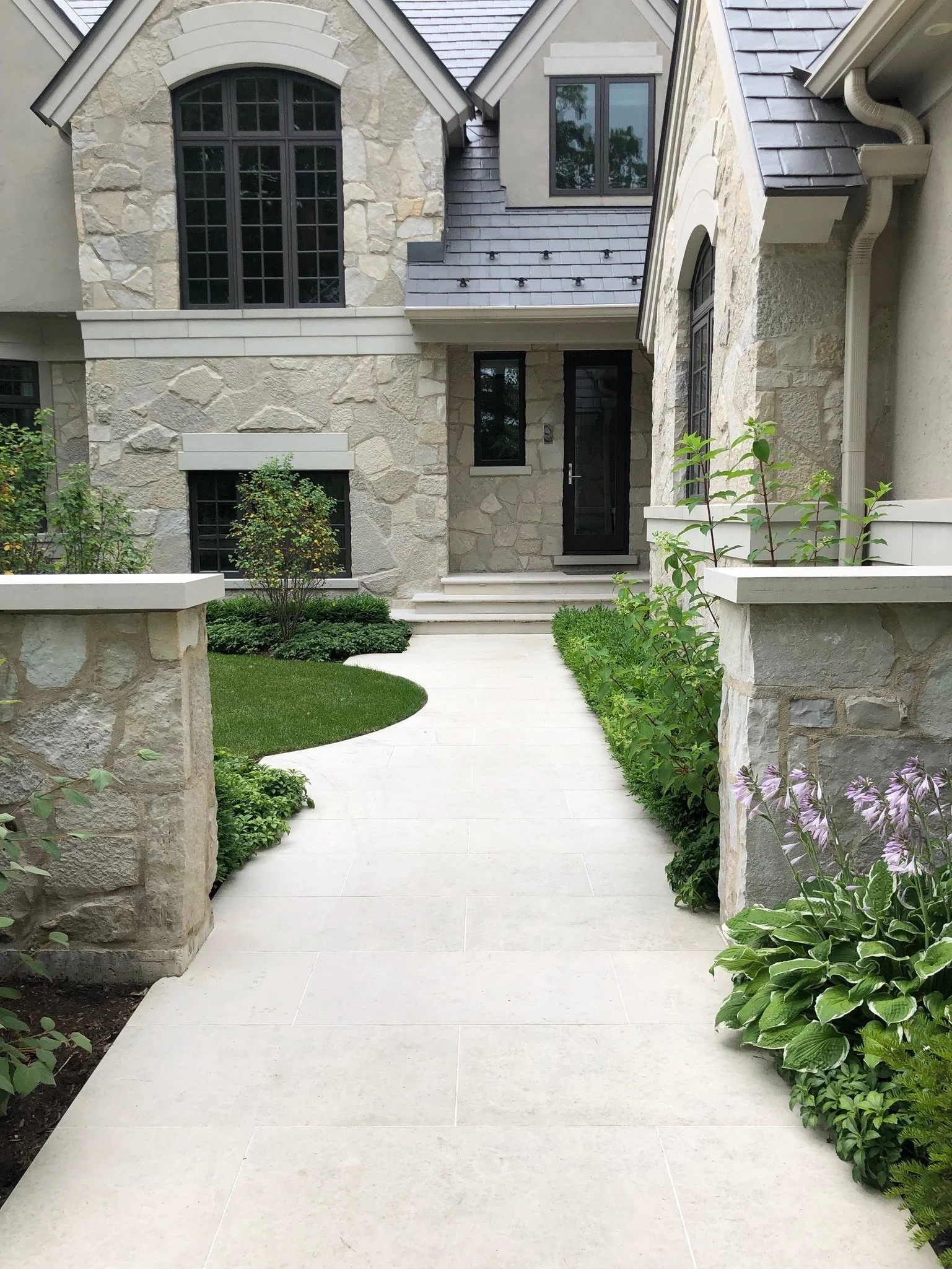 A stone house with large windows, a gray roof, and a small front yard with a porch, steps, and greenery.