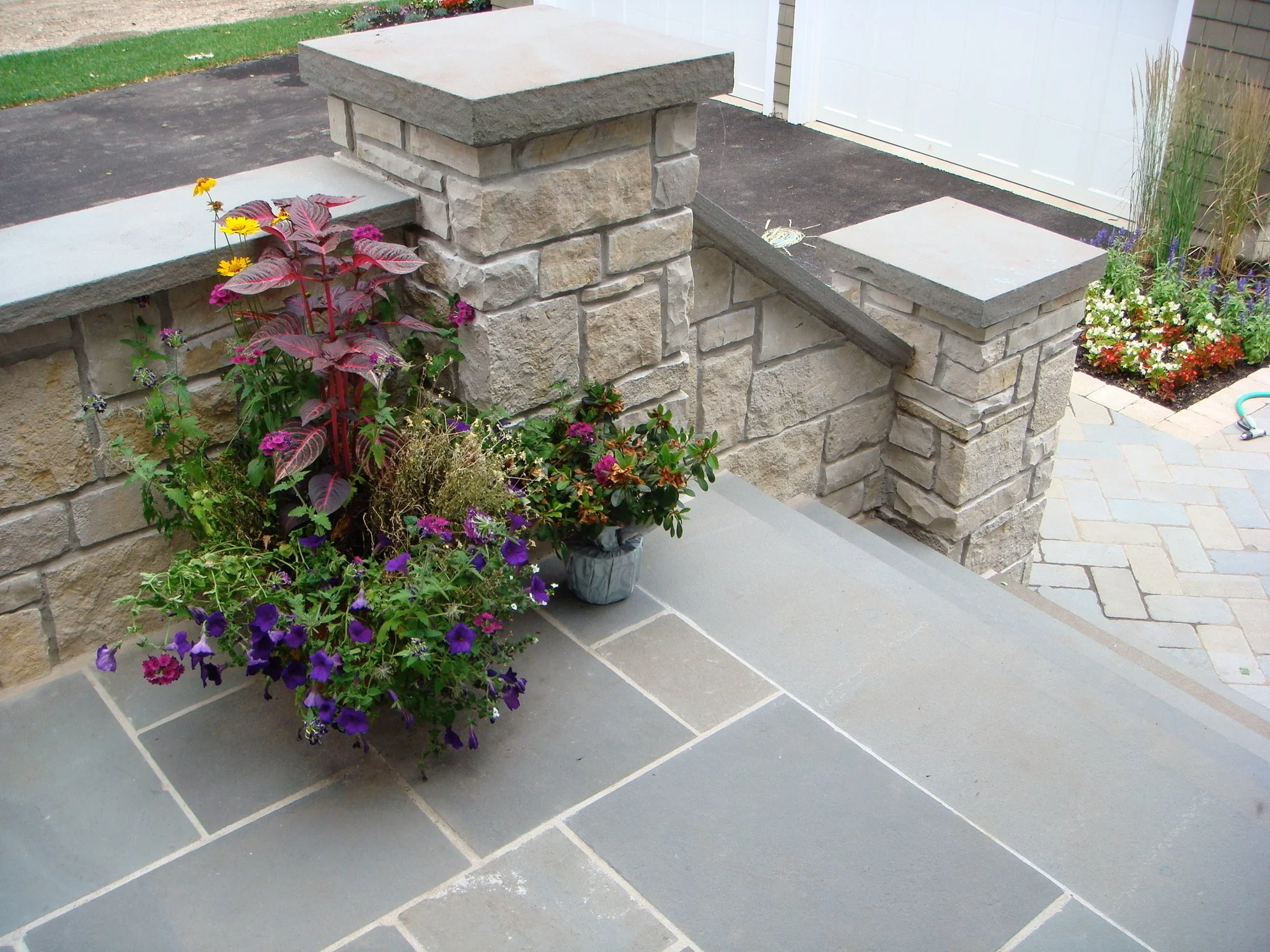 A stone wall with a flat stone cap on top, next to a flower bed with colorful flowers, and a concrete walkway leading to a white door.