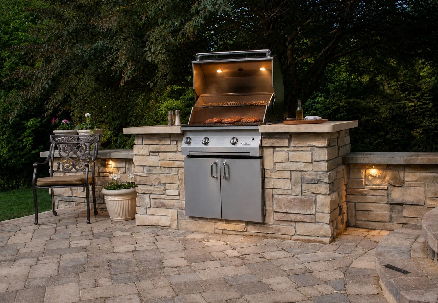 An outdoor stone patio with a built-in gas grill that has food cooking inside, illuminated by built-in lights. To the left, there is a decorative metal chair with a cushion and a potted plant. The area is surrounded by trees and greenery.