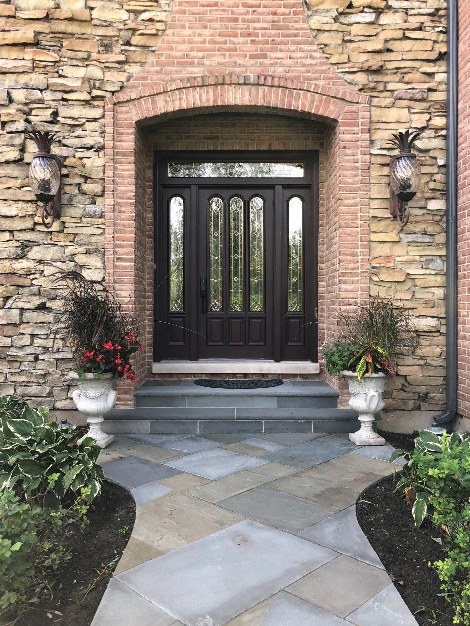 Front entrance of a house with a black door with glass panels, surrounded by stone and brick wall, two wall-mounted lanterns, and flower pots with plants and flowers on each side of the door.