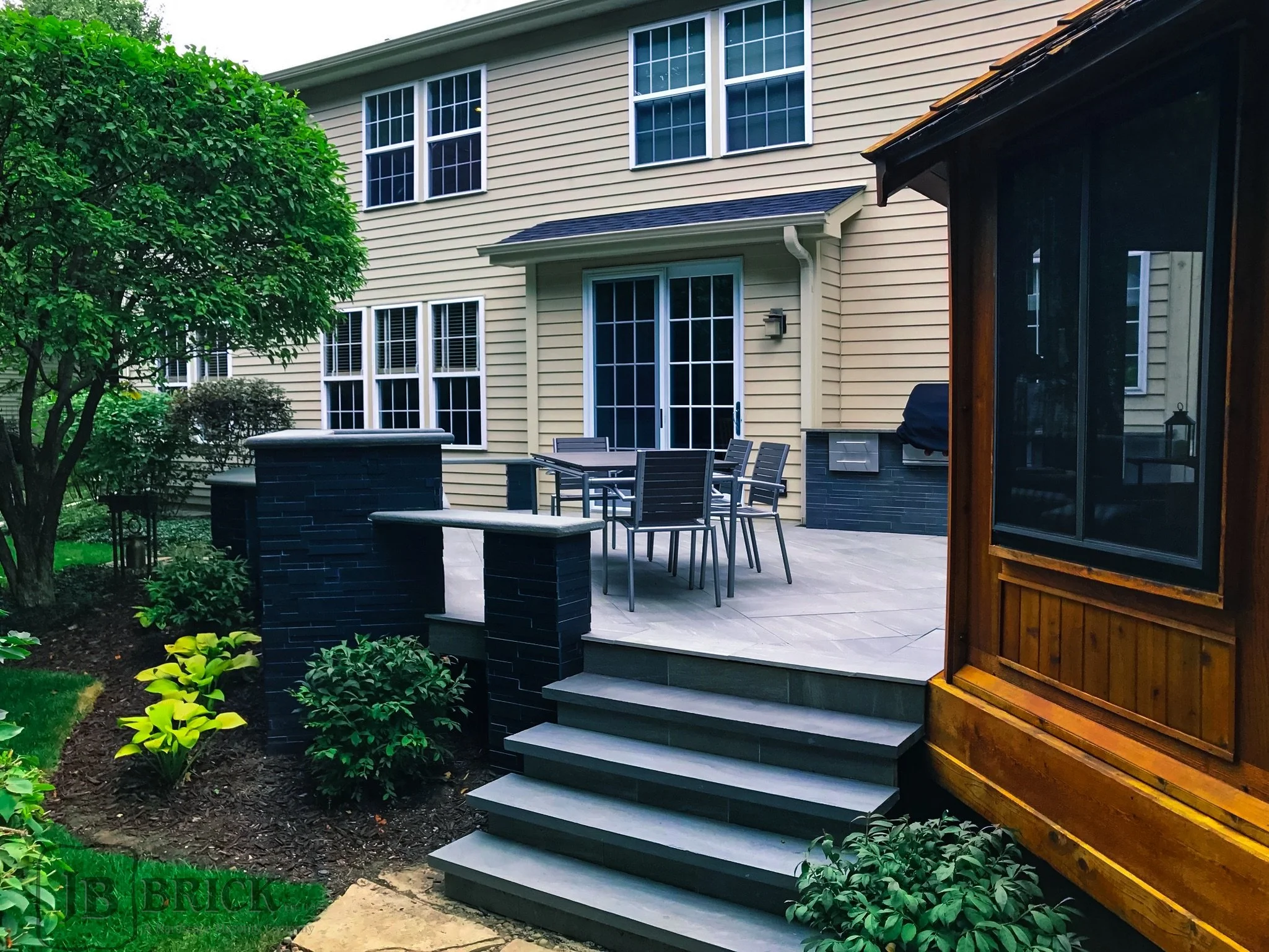 Backyard patio with outdoor dining table, chairs, and stairs leading down to garden area, with a house in the background and greenery around.