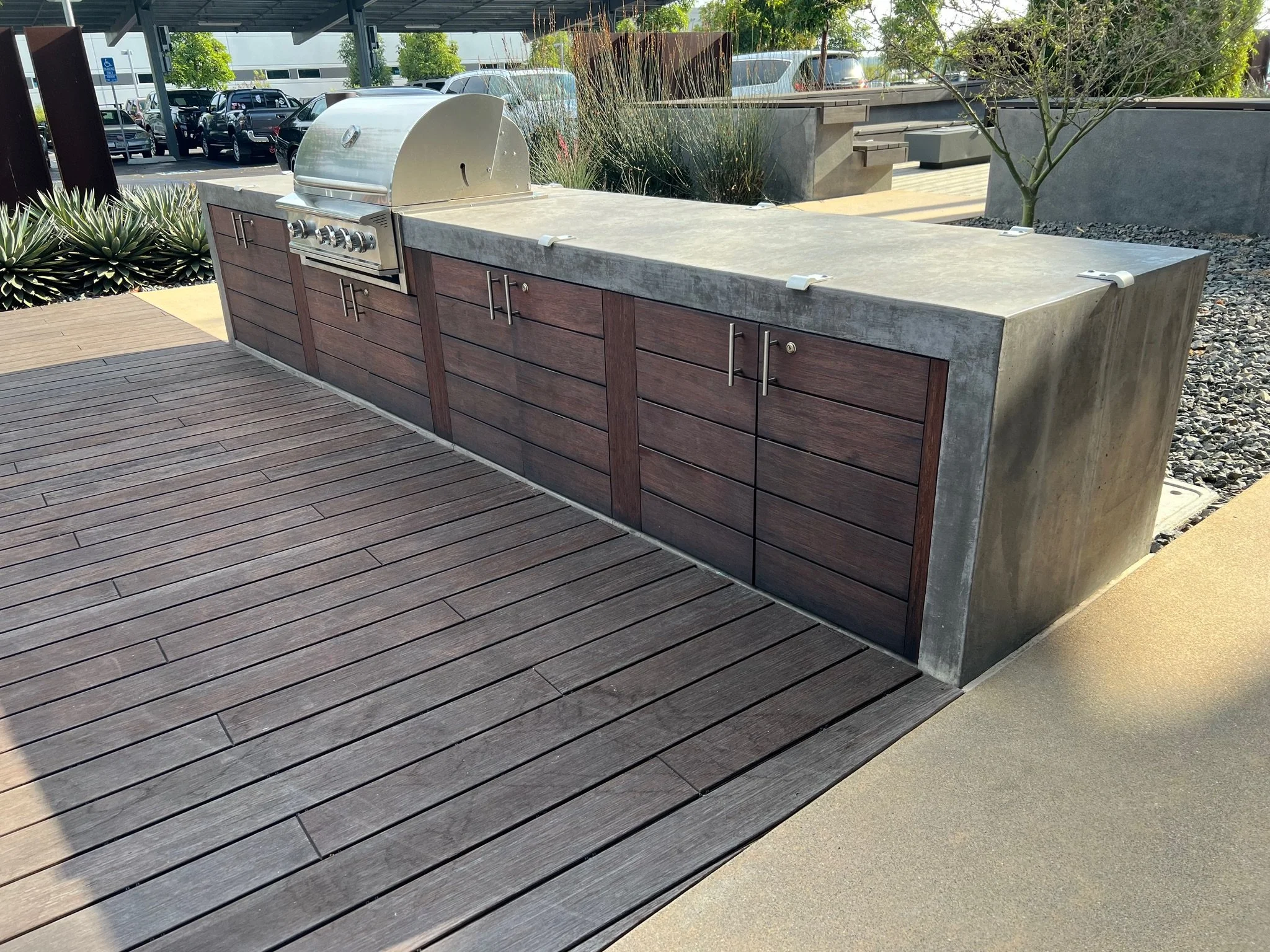 Outdoor community barbecue area with a stainless steel grill, surrounded by wooden cabinets and a concrete countertop, with landscaping including plants, trees, and a parking lot in the background.