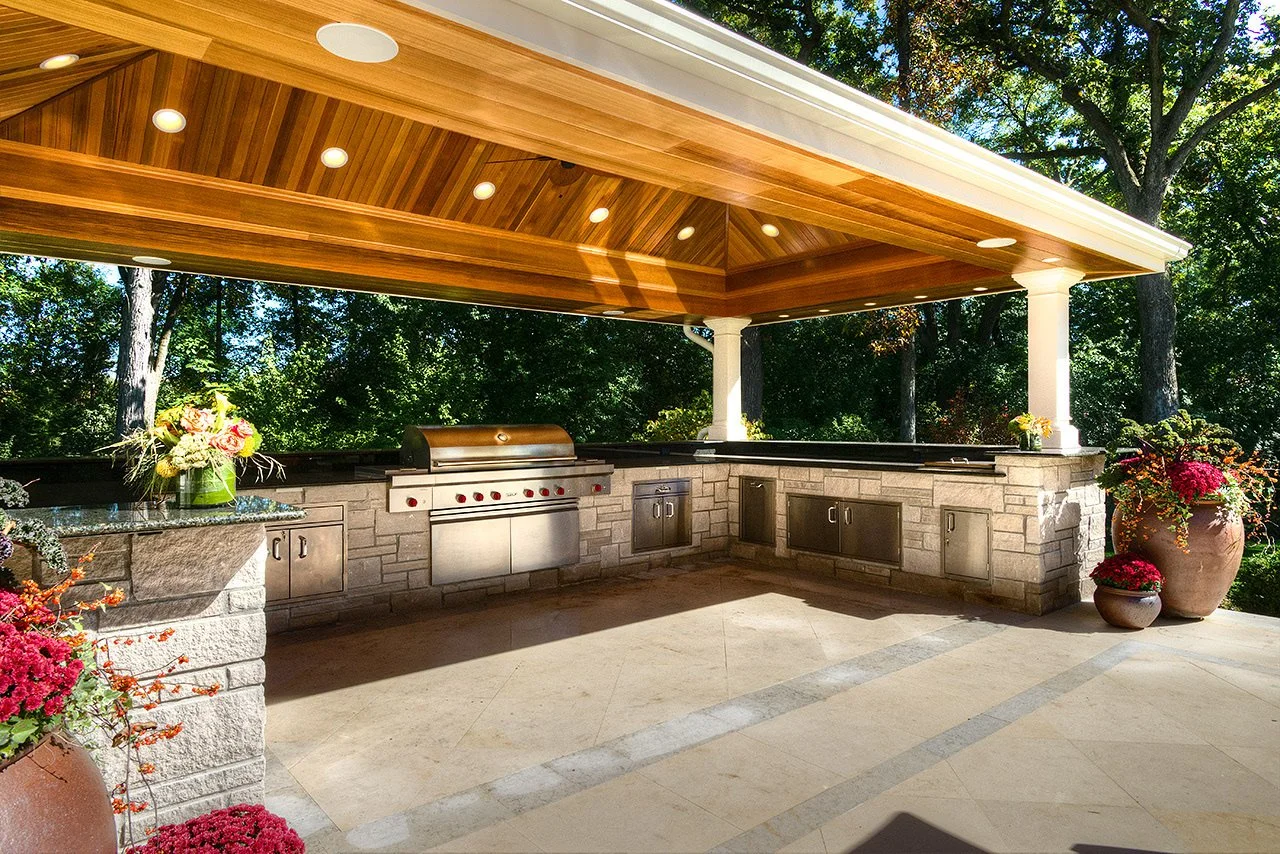Outdoor kitchen with built-in grill, stone counters, and wooden ceiling, surrounded by trees and decorative pots with flowers.