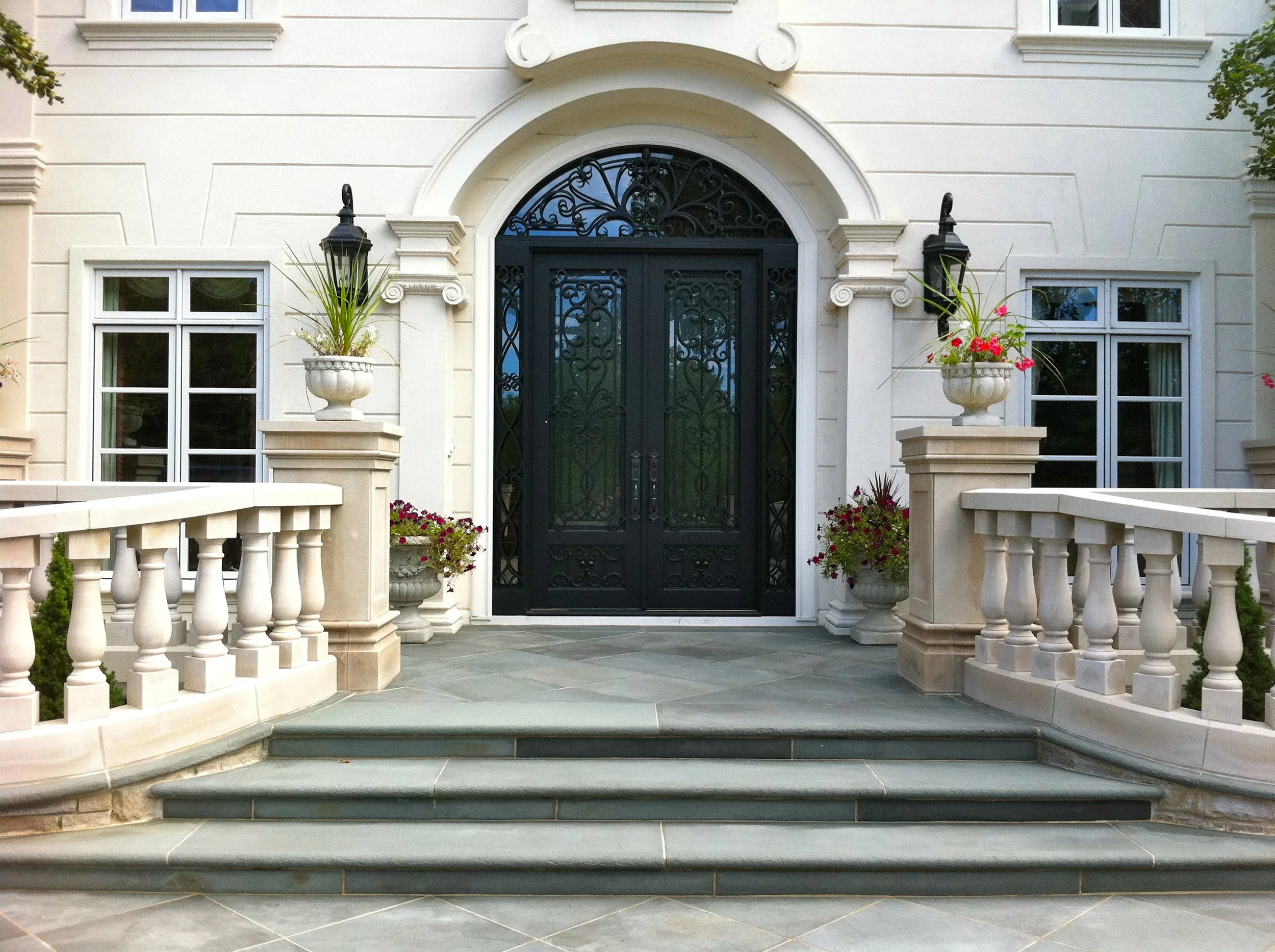 Front entrance of a white building with a black wrought iron gate, flanked by two wall-mounted lanterns, and decorative moldings around the windows and door.