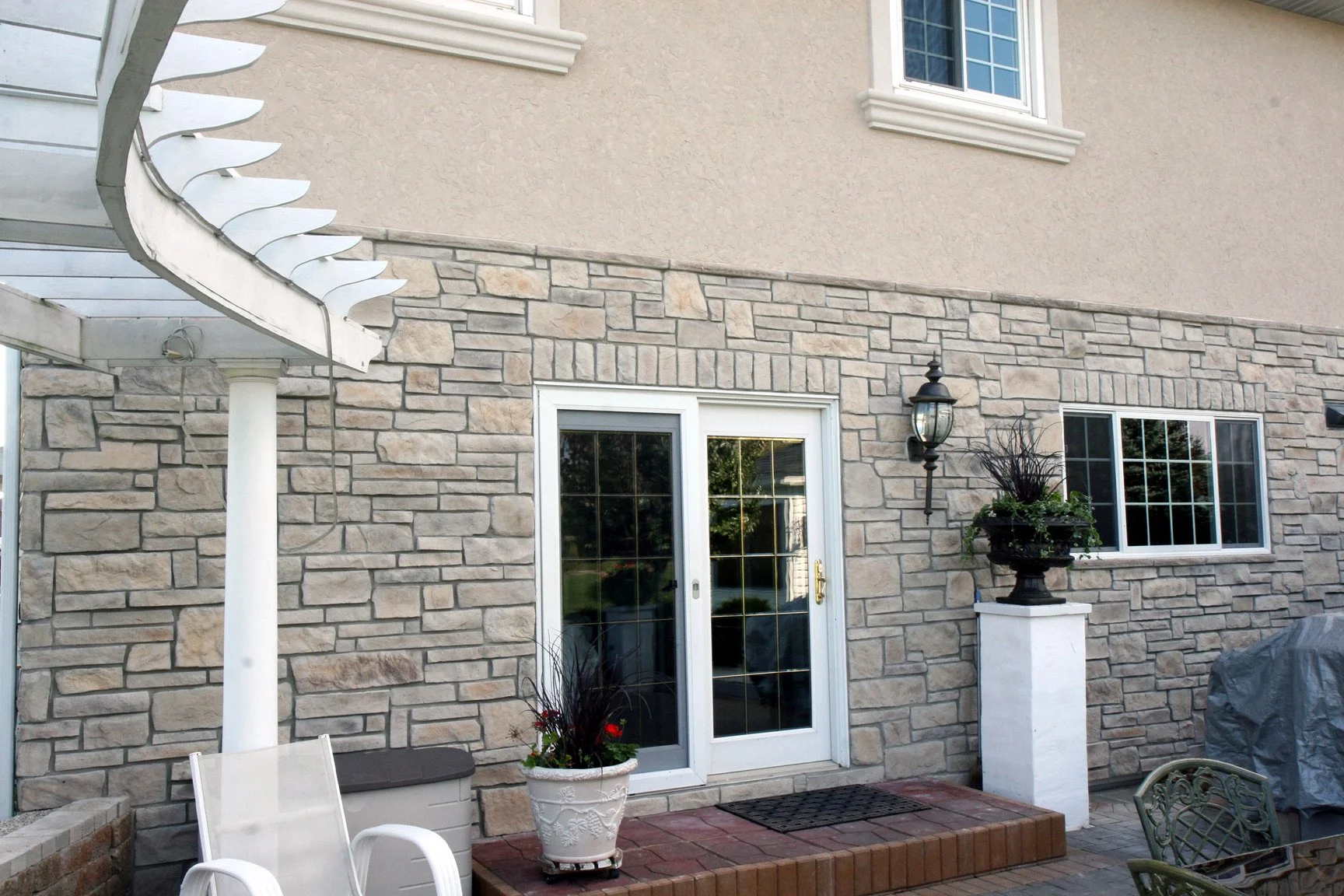 Backyard patio area with stone and stucco wall, sliding glass door, flower pots, outdoor furniture, lantern-style wall light, and grill covered with a cover.