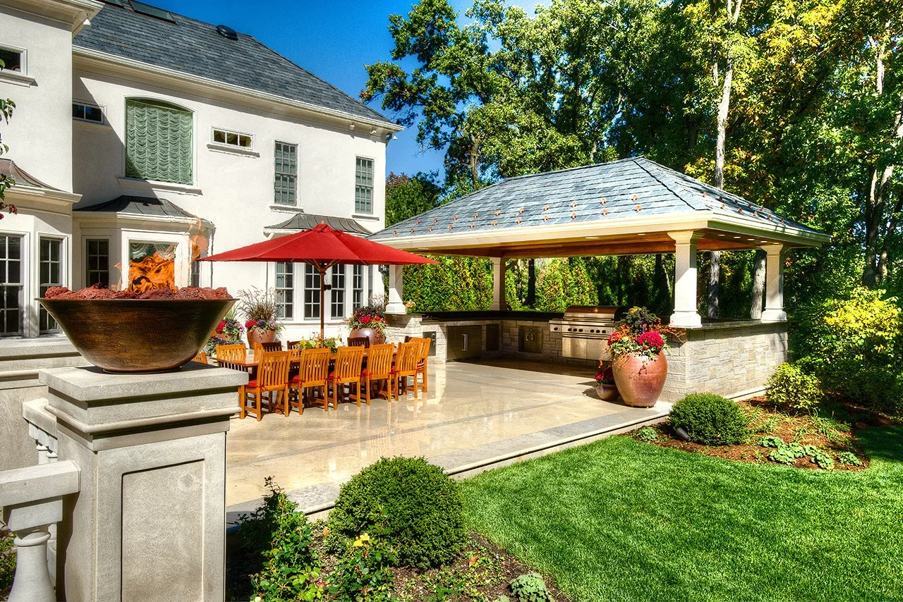 Backyard patio area with a large bowl-shaped fire feature, a wooden dining table with an orange umbrella, and a covered outdoor kitchen with grill and stone counter, surrounded by greenery and trees.