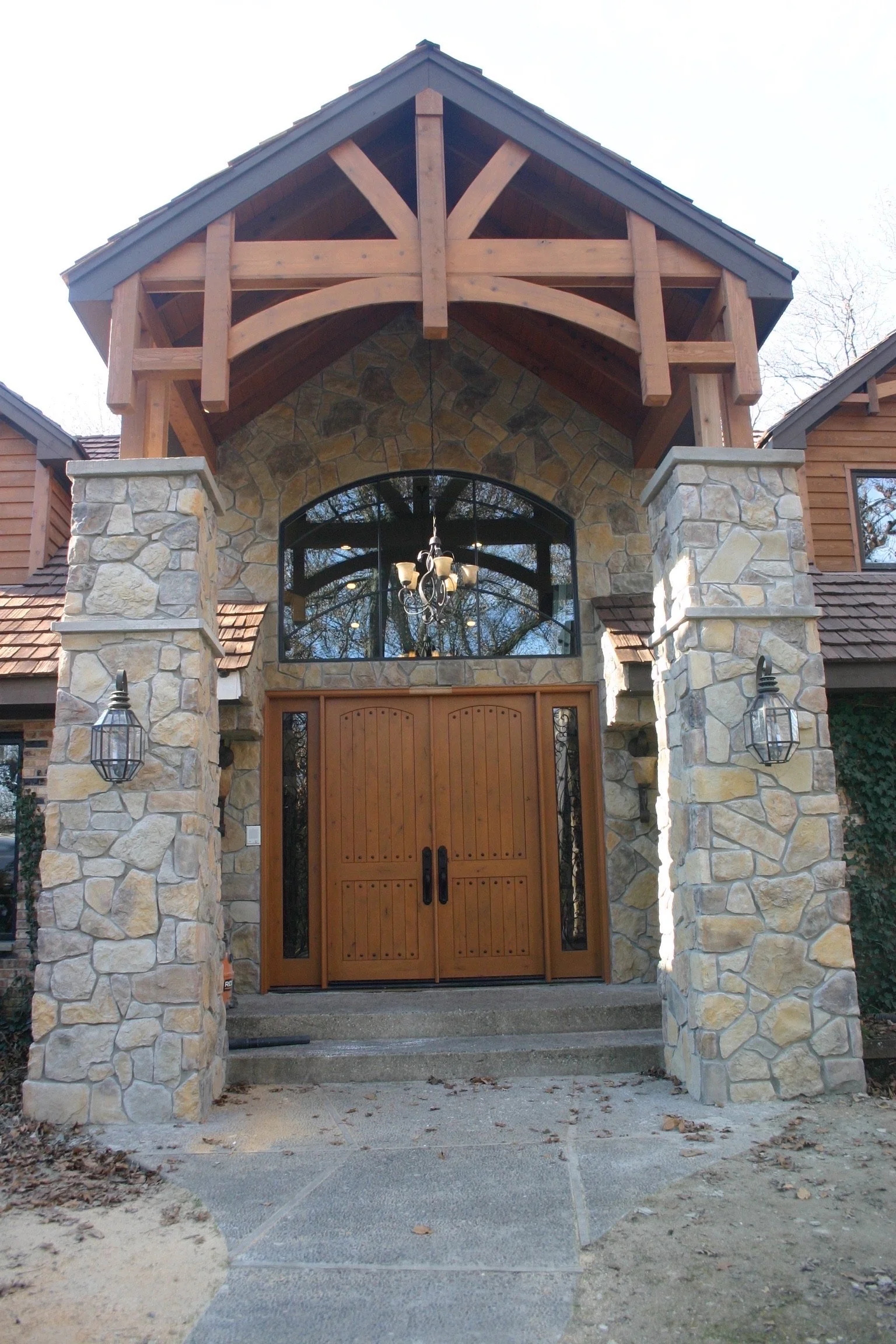 Stone and wood front entrance with wooden double doors, stone pillars, decorative lanterns, and an arched window above the door, featuring a wooden gable roof with support beams.