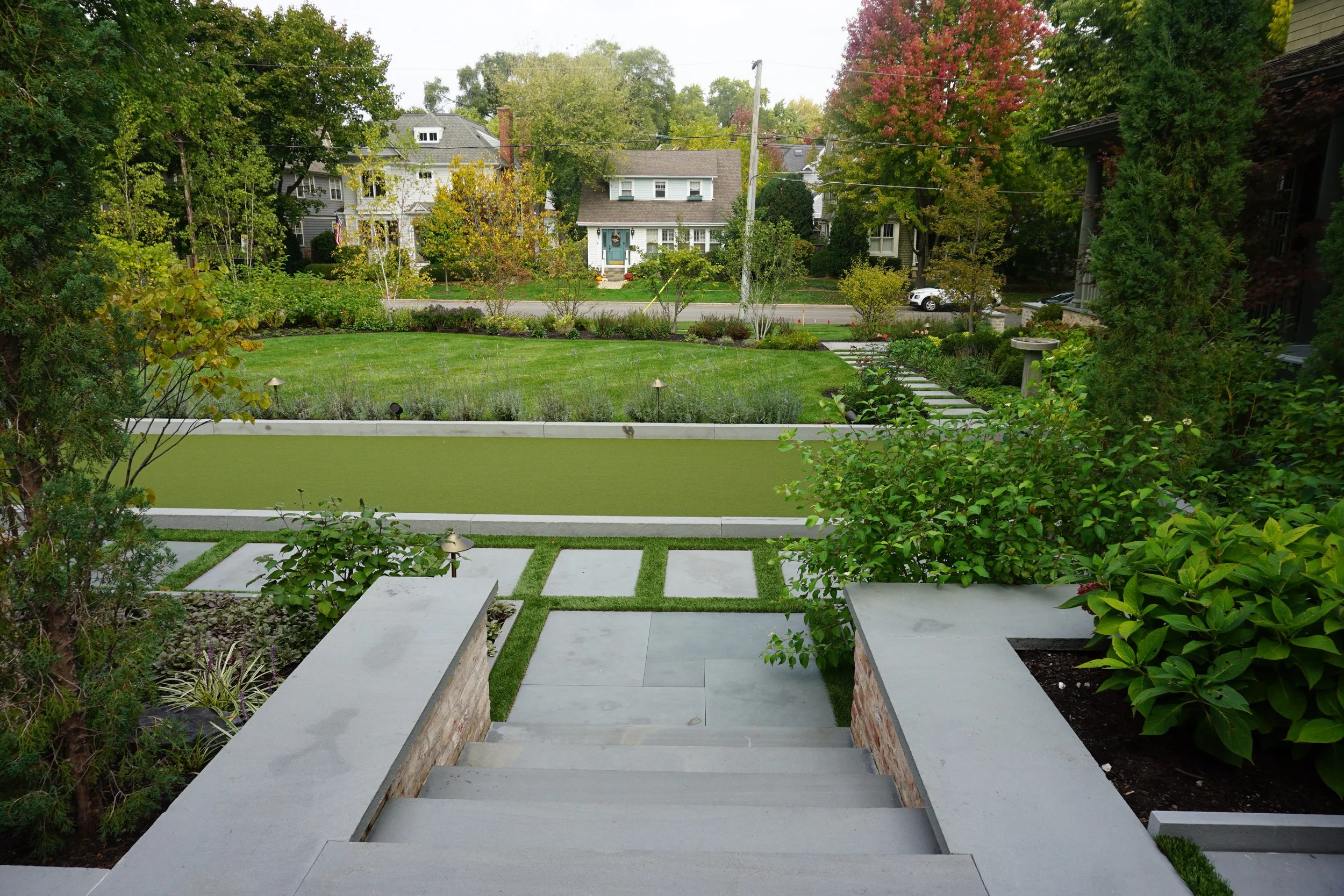 View of a backyard garden with steps leading down from a porch, green grass, shrubs, and trees with autumn foliage, and a street with houses and parked cars in the background.