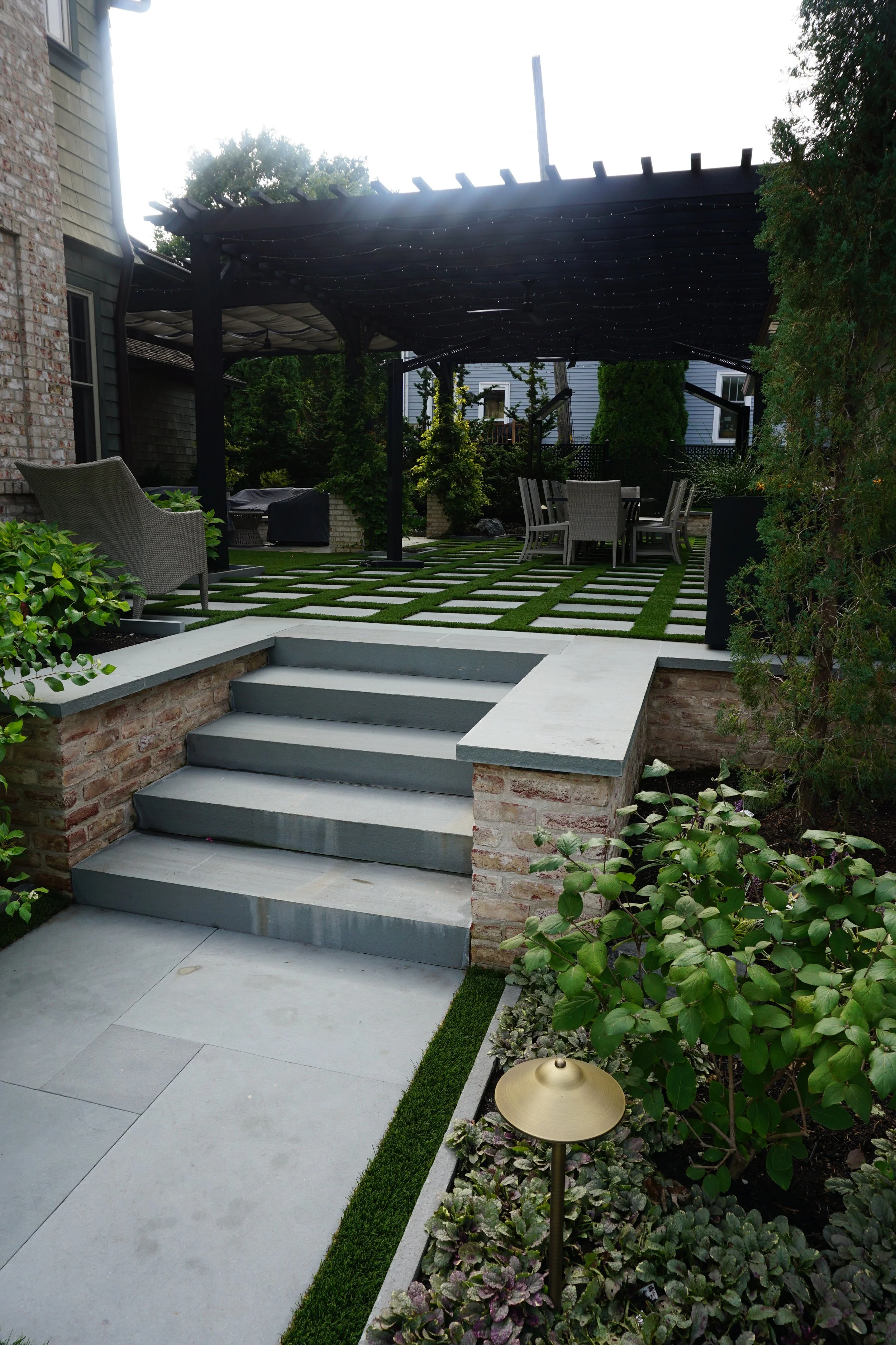 A backyard patio with gray steps leading to a paved area, surrounded by lush green plants and a small garden area with low-growing plants. There is a black pergola overhead with string lights, and outdoor furniture including a round table with chairs