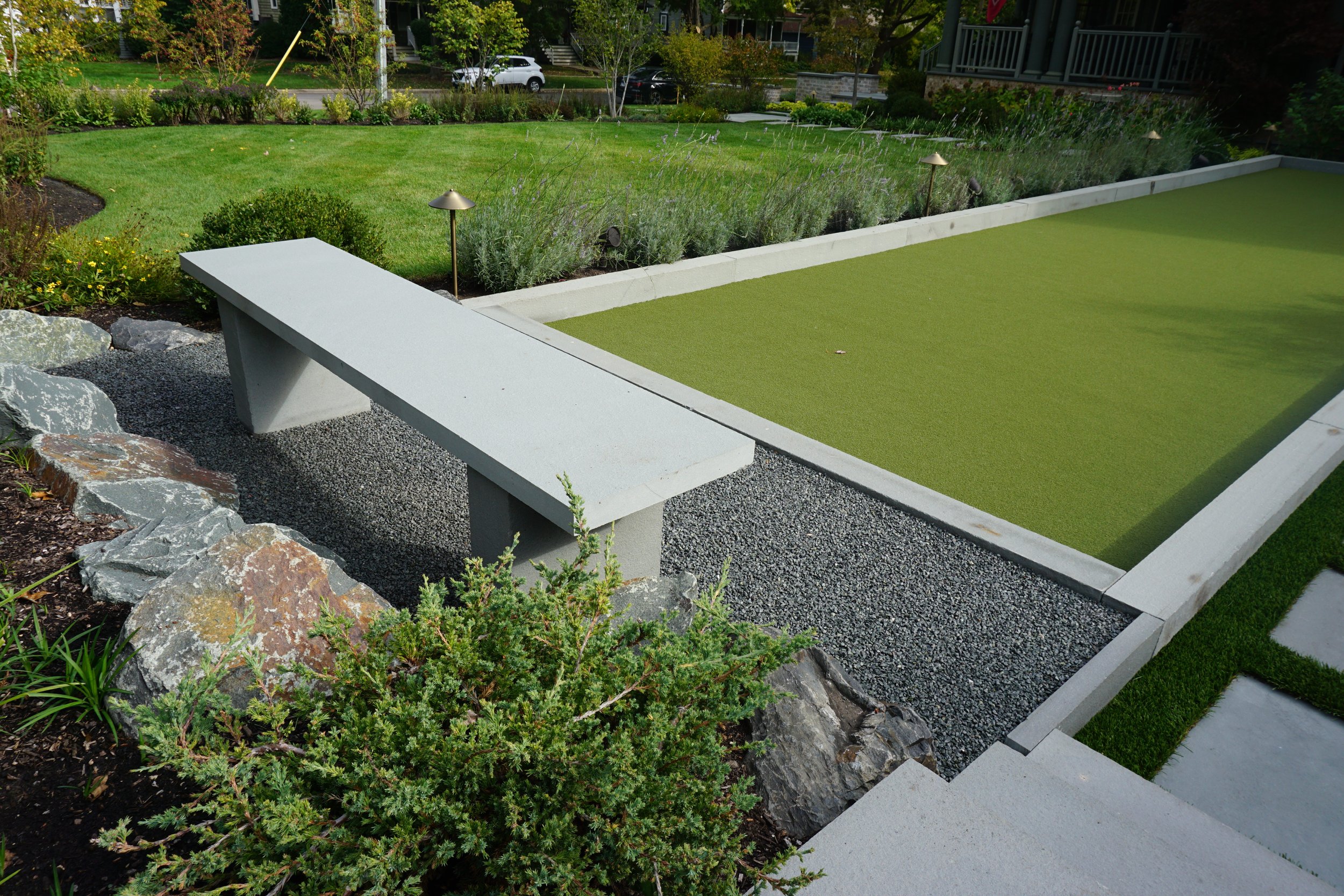 A backyard with a well-maintained grassy lawn and a border of plants and shrubs. There is a concrete or stone patio with a green artificial turf square, surrounded by a stone and gravel landscape. Small pathway lights are along the border of the planting bed.