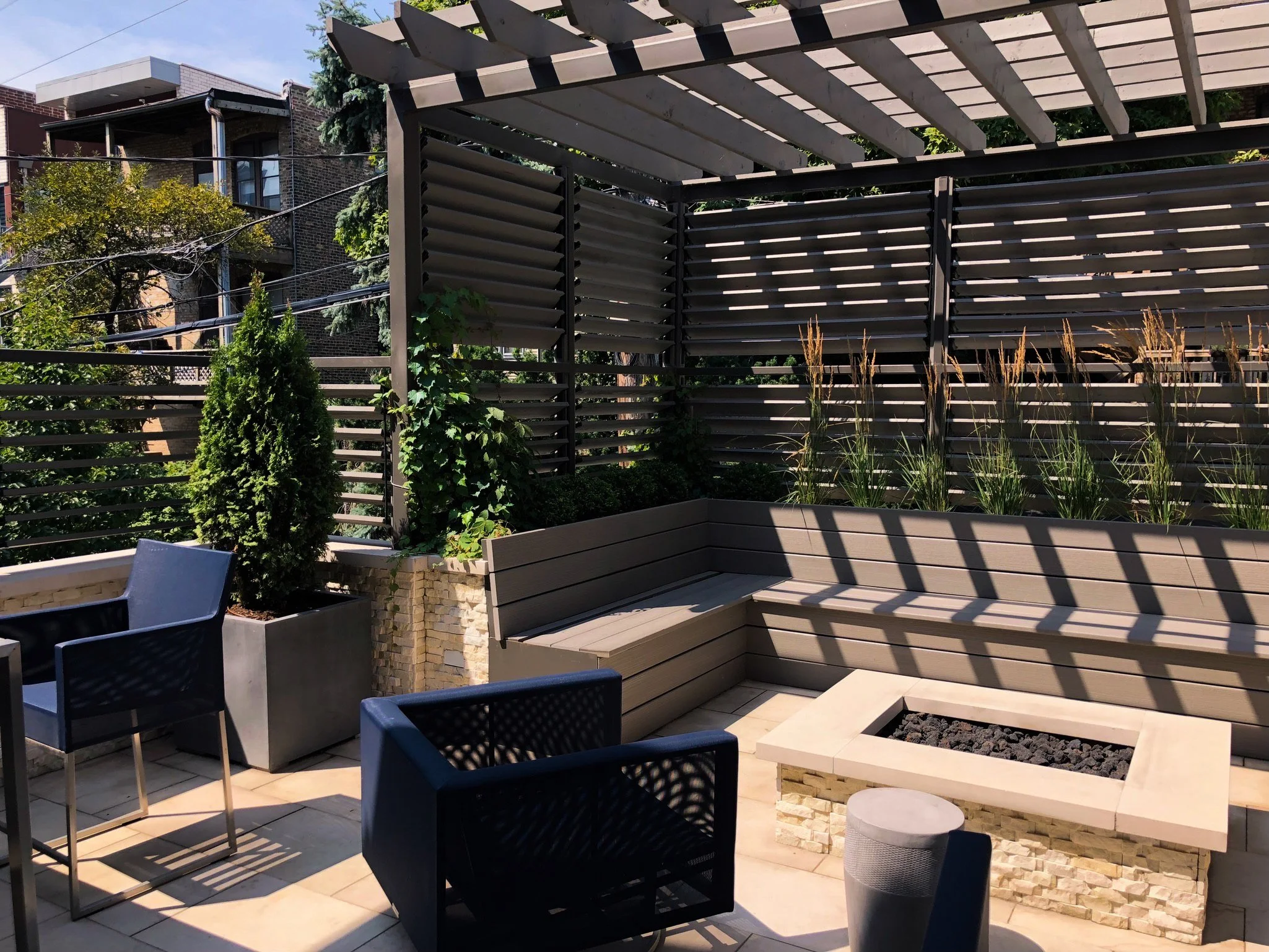 Modern outdoor patio with black chairs, a built-in fire pit, and a privacy fence made of horizontal wood slats, with trees and residential buildings in the background.