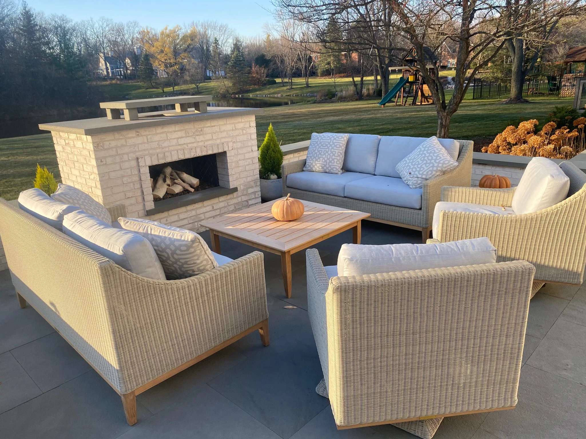 Outdoor patio with wicker sofa and chairs, a small wooden table with pumpkins, a brick fireplace, and a view of a grassy yard with trees, a pond, and houses in the background.