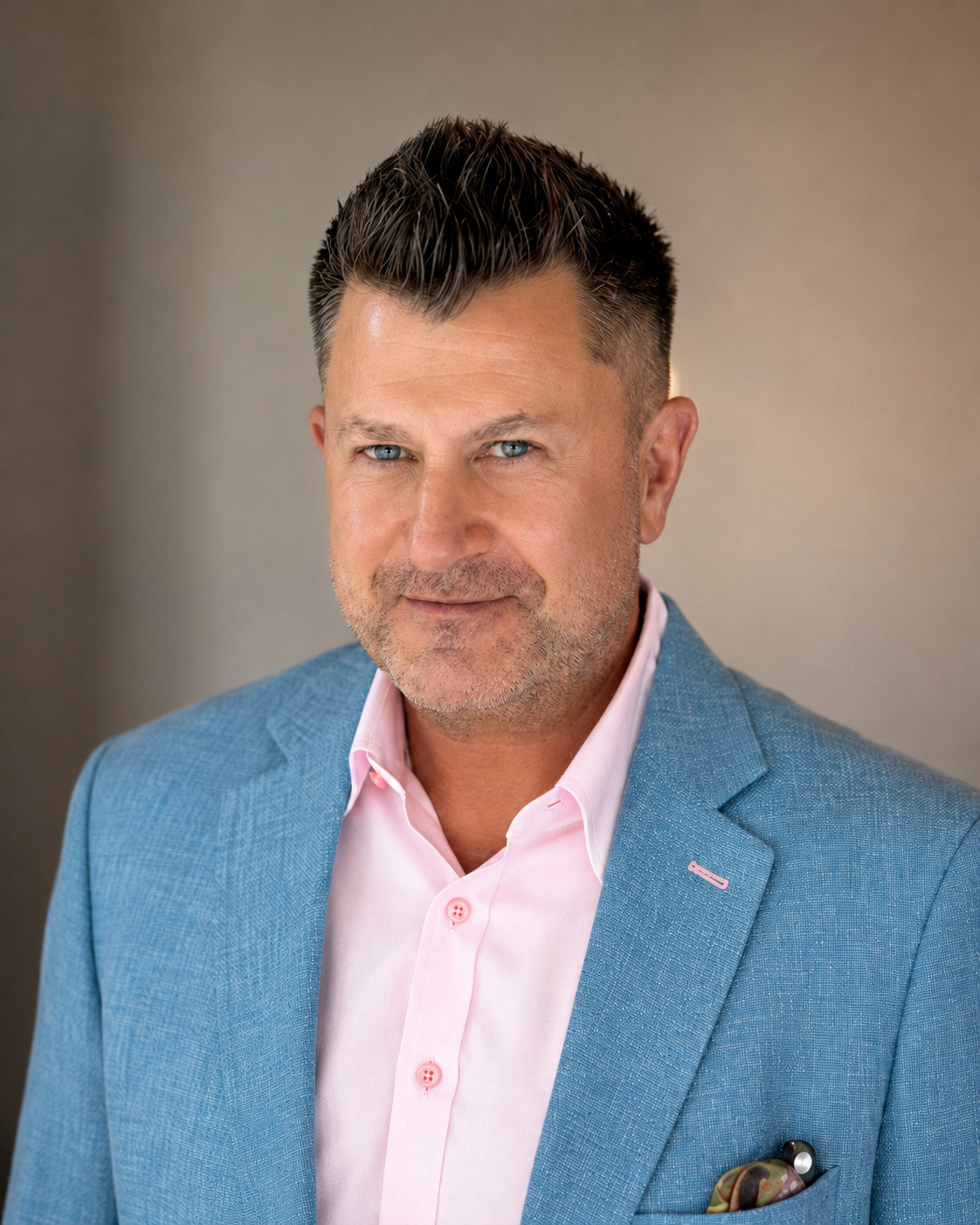 Headshot of a middle-aged man with blue eyes, short dark hair, and stubble, wearing a light blue blazer over a pink dress shirt, with a pocket square and pen in his blazer pocket, against a neutral background.