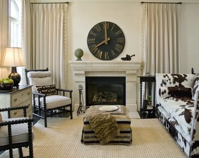 Living room with fireplace, large wall clock, cowhide-patterned sofa, black side table, armchair with pillow, and curtains.