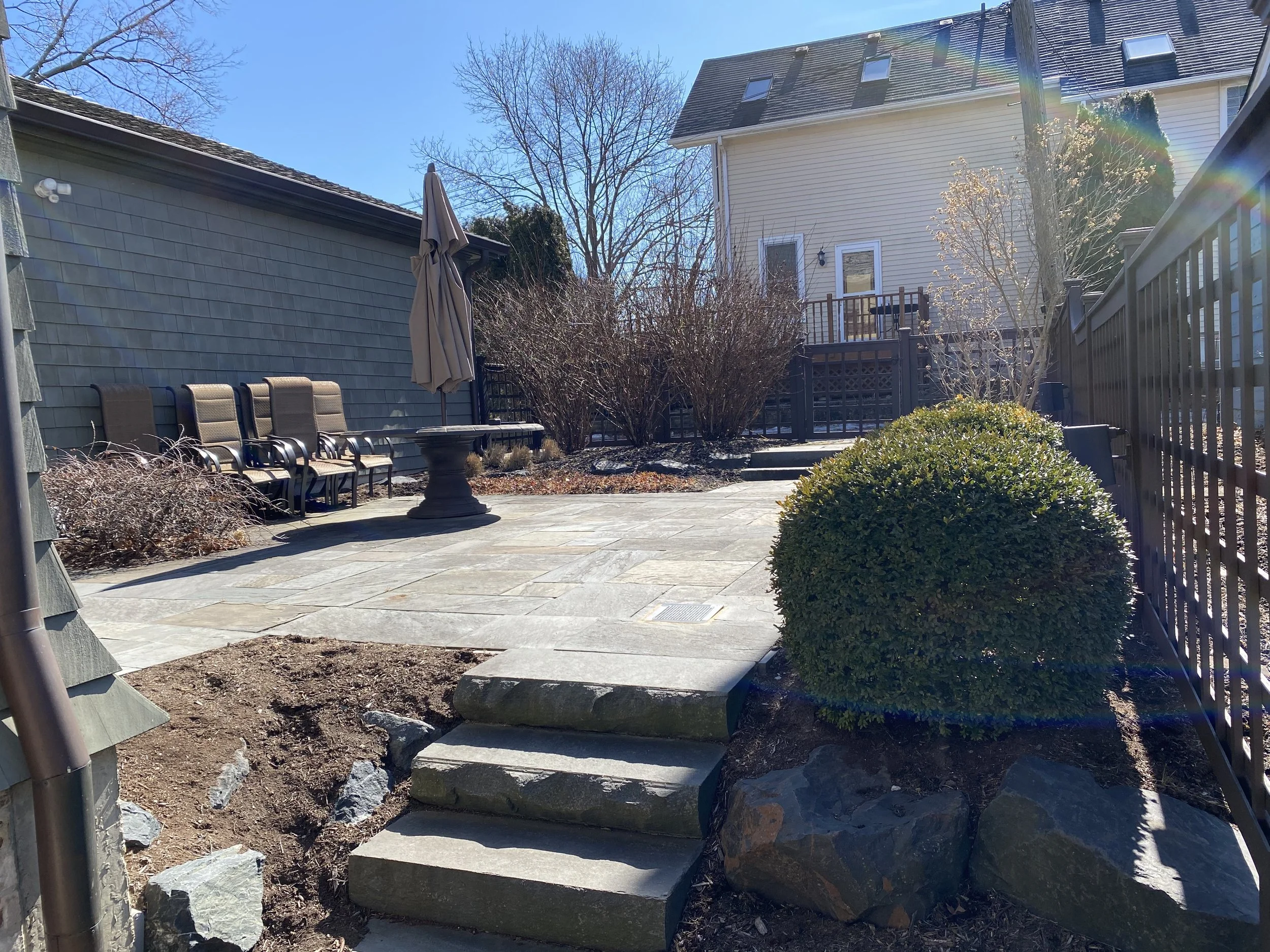 A backyard patio area with stone steps, a fenced yard, trees without leaves, a building with light yellow siding, and patio chairs with a closed umbrella.