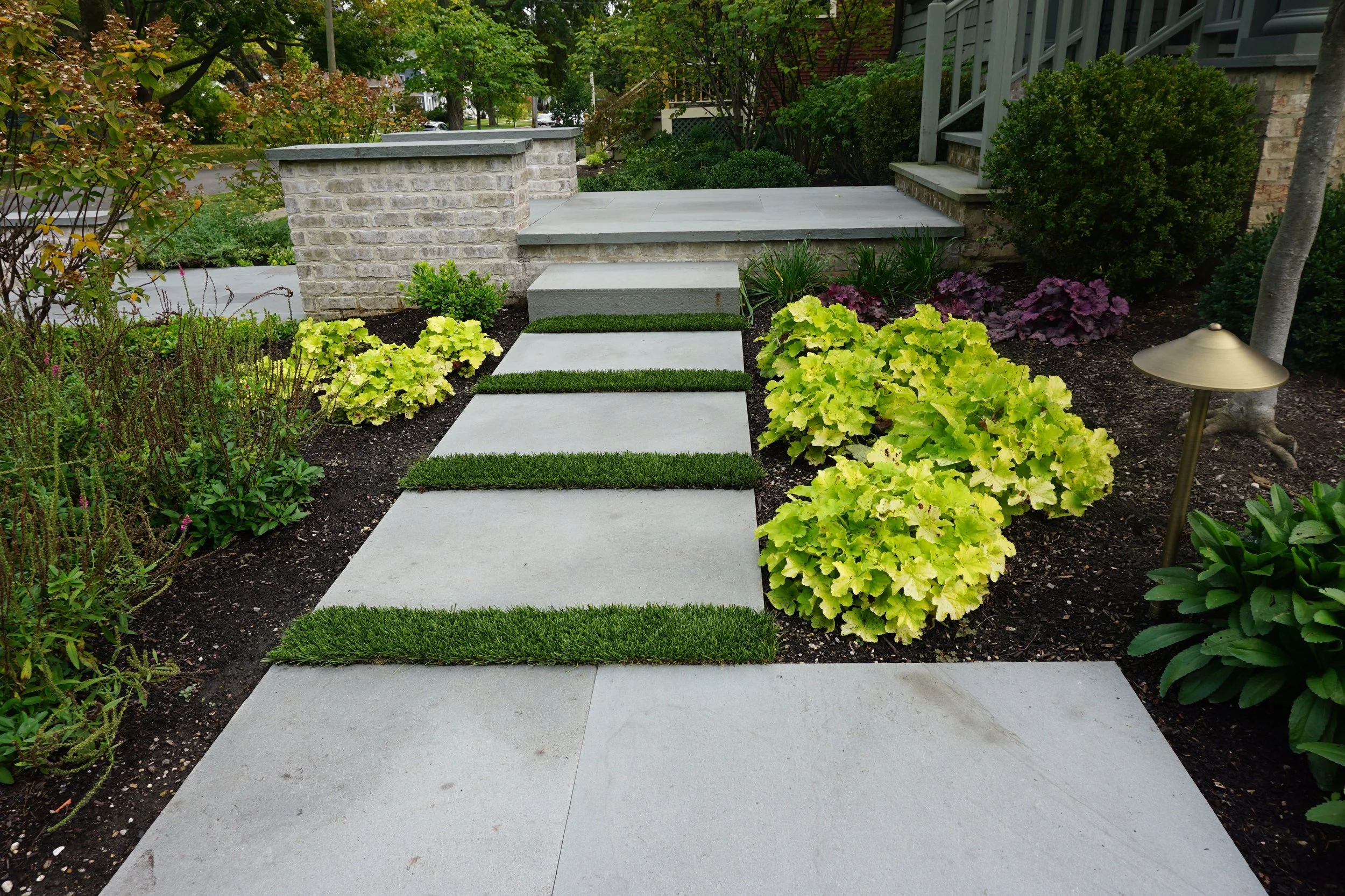 Front yard with concrete steps and walkway, surrounded by yellow and purple flowering plants and bushes, with a lamp and trees lining the street in the background.