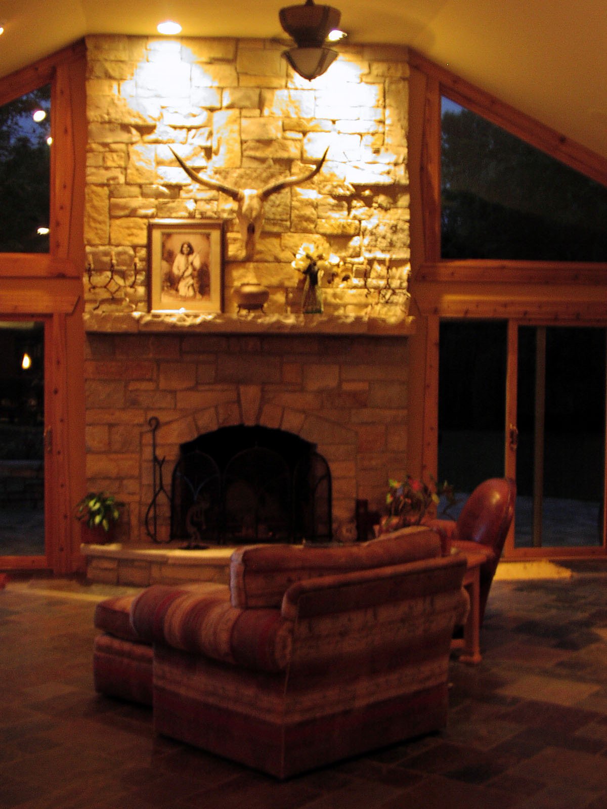 A cozy living room with a stone fireplace, decorated with a framed religious picture, a deer skull with antlers, and floral arrangements on the mantel. Plush armchairs and large windows create a warm atmosphere.