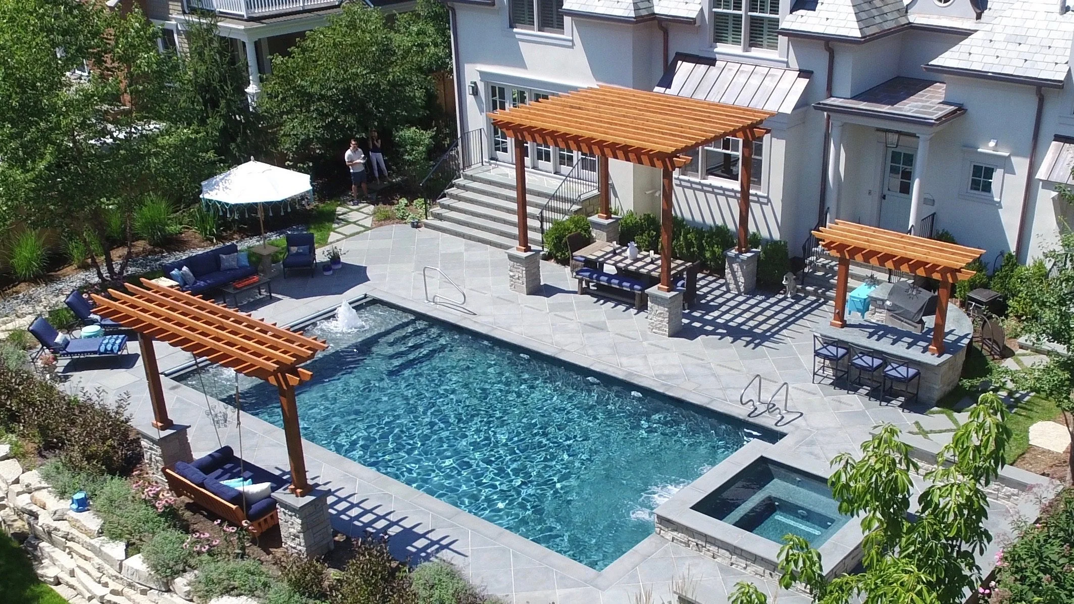 A backyard swimming pool area with shade structures, outdoor furniture, and lush greenery.