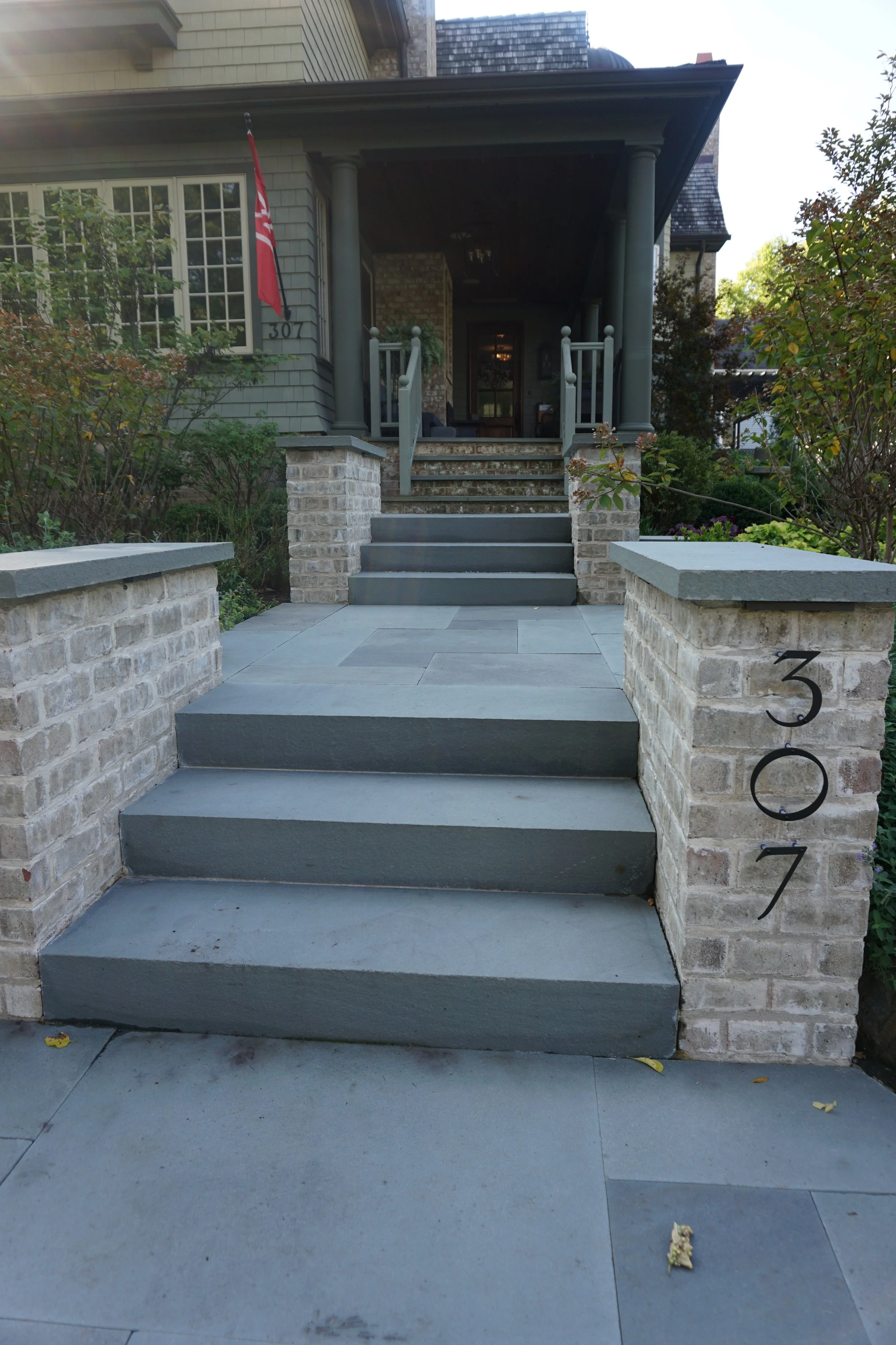 Front porch of a house with stairs, brick columns, plants, and a red flag. The house number is 307, and the patio has a porch with a chandelier visible inside.