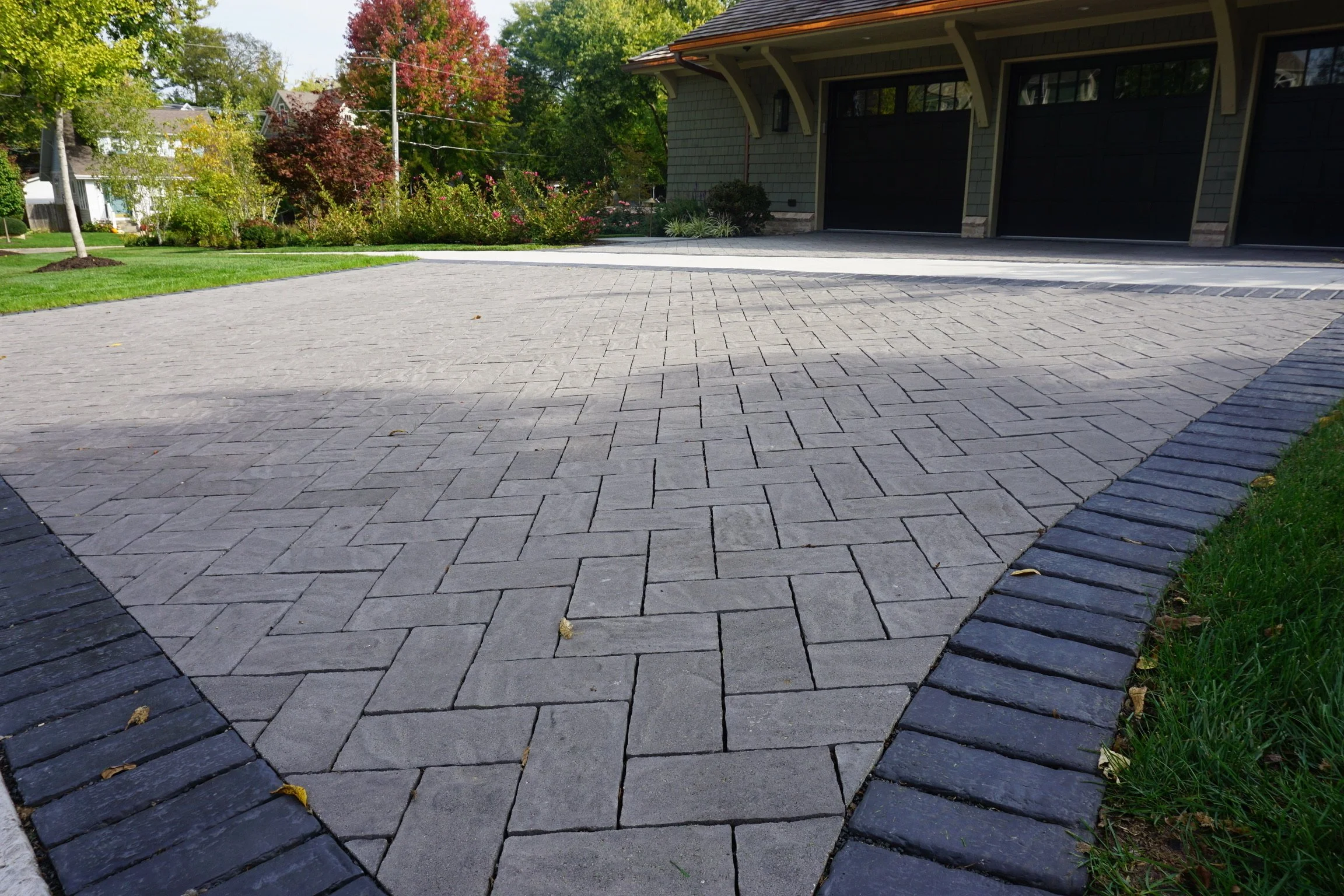 A residential driveway made of interlocking gray stone pavers, with a curved black border along the edges, leading to a house with a garage. The house has dark-colored garage doors, siding, and architectural details. In the background, there are lush