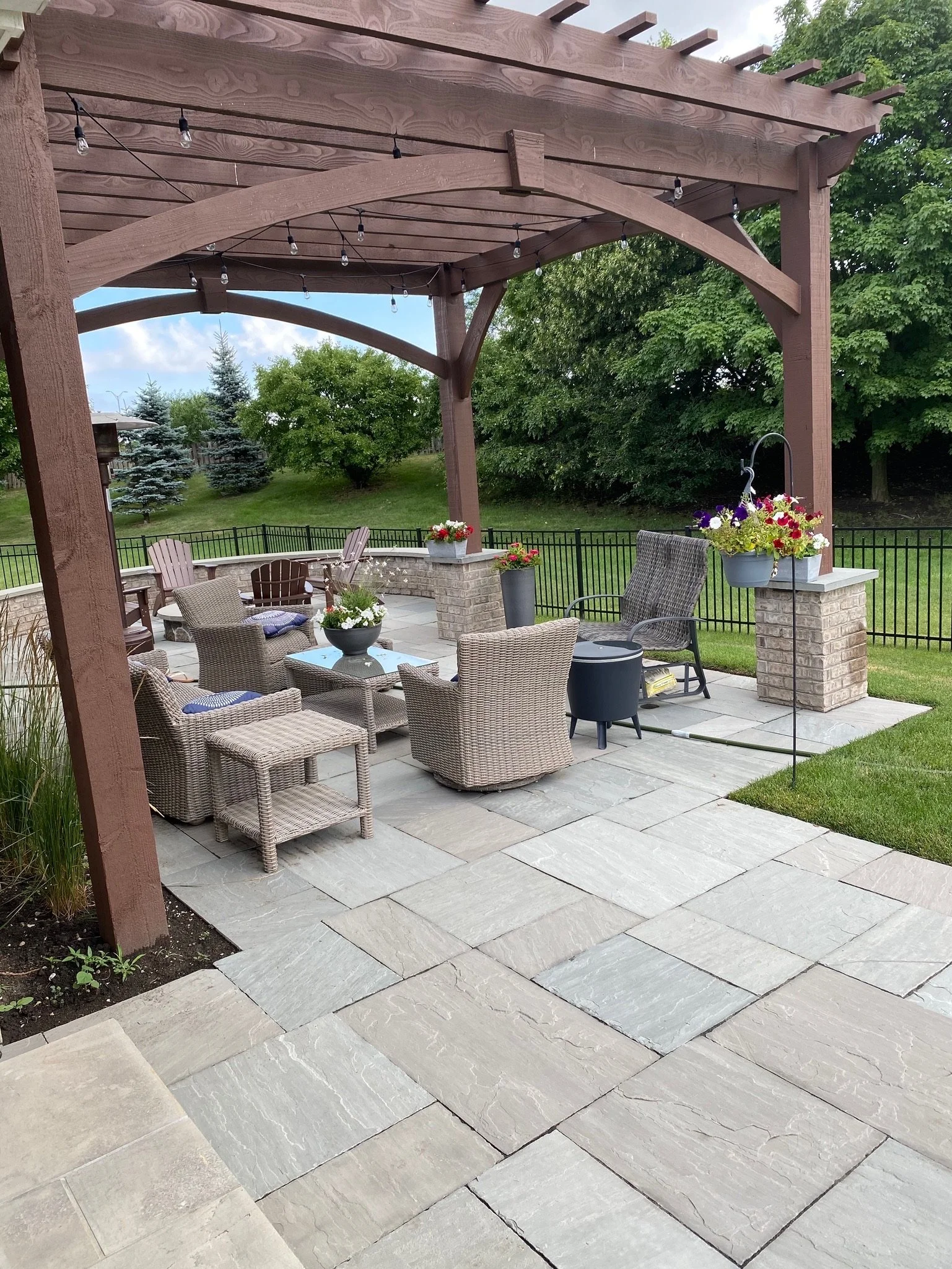 A backyard patio with sitting area, decorated with potted flowers, surrounded by a metal fence, with trees and greenery in the background.
