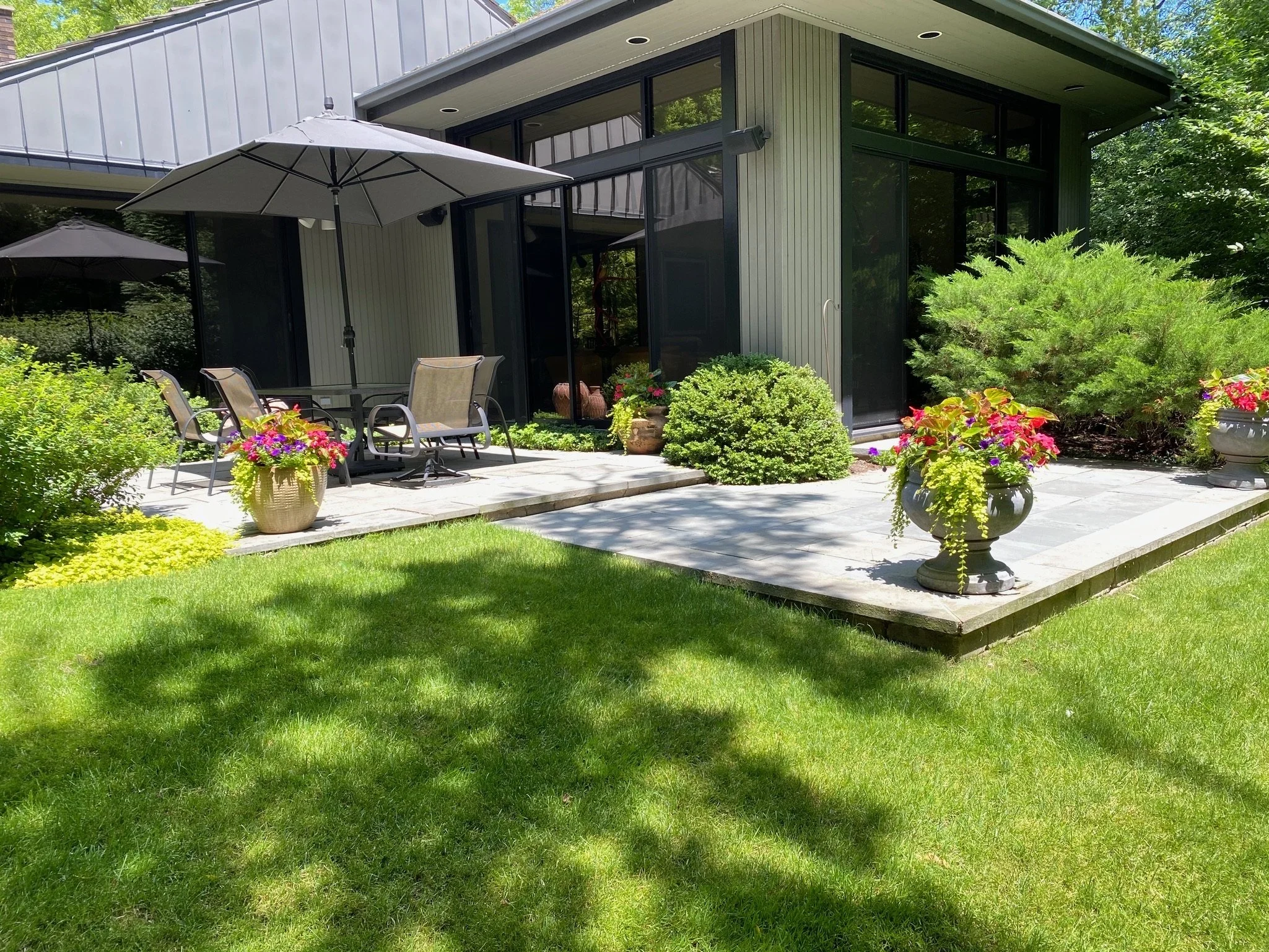 An outdoor patio area adjacent to a modern house with large glass windows and sliding doors. The patio has a table with umbrellas, chairs, and various potted plants and flower arrangements. The area is surrounded by greenery, including bushes and a l