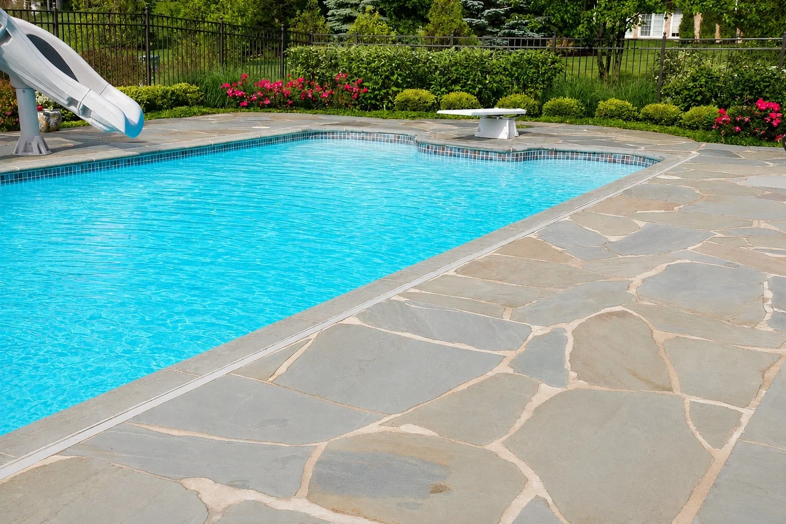 A backyard swimming pool with clear blue water, surrounded by a stone patio, flower beds with pink and yellow flowers, greenery, and a black metal fence.