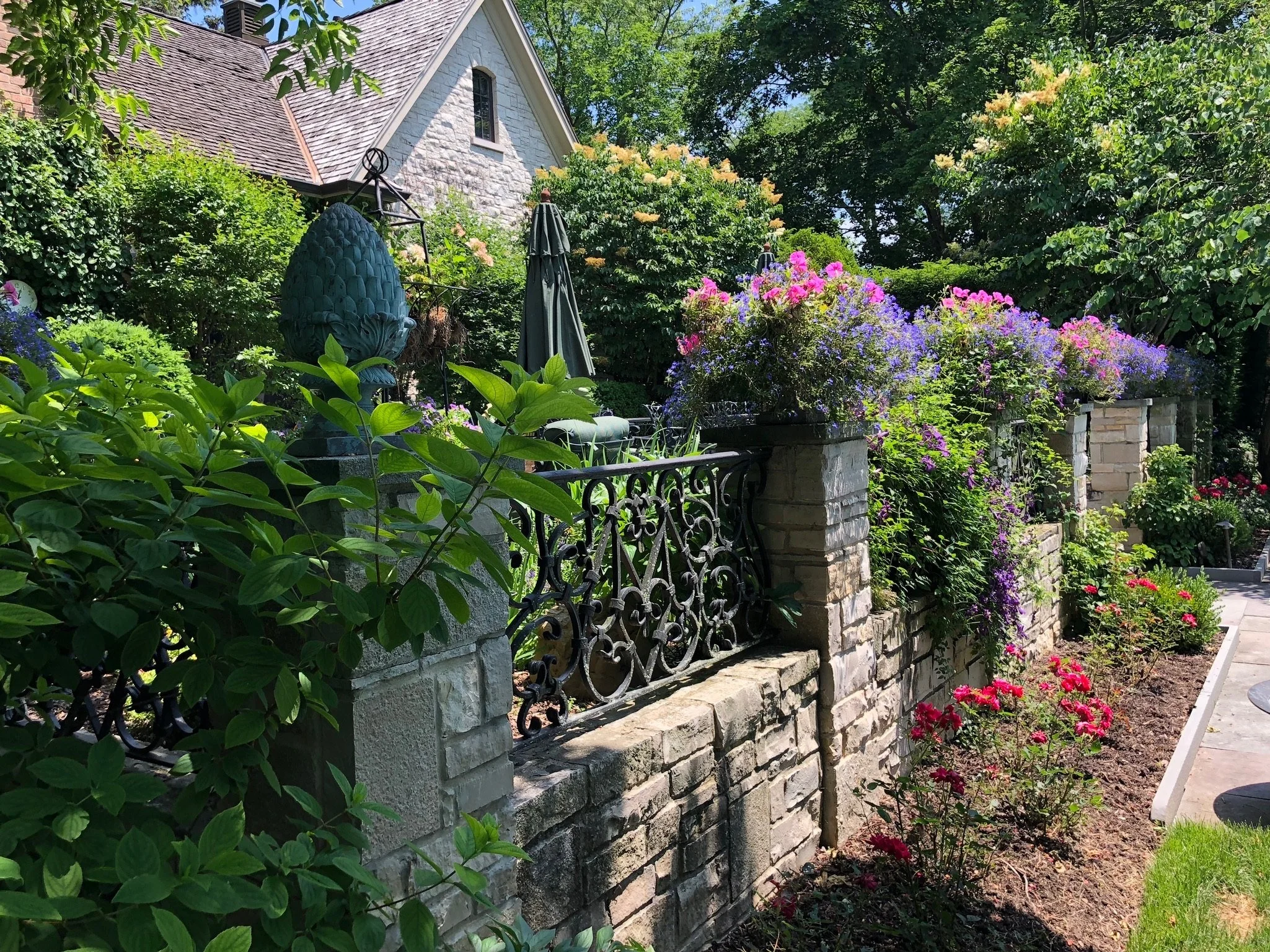 A garden with a stone wall, metal fence, and colorful flowers including pink, purple, and red, with a house in the background and trees surrounding the area.