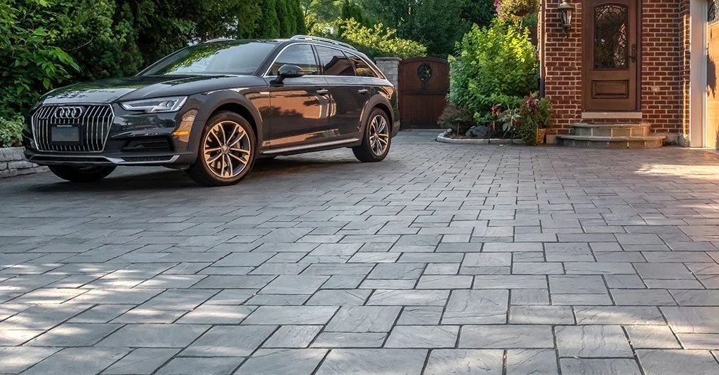 A black Audi station wagon parked on a paved driveway in front of a brick house with steps and a wooden door, surrounded by greenery.