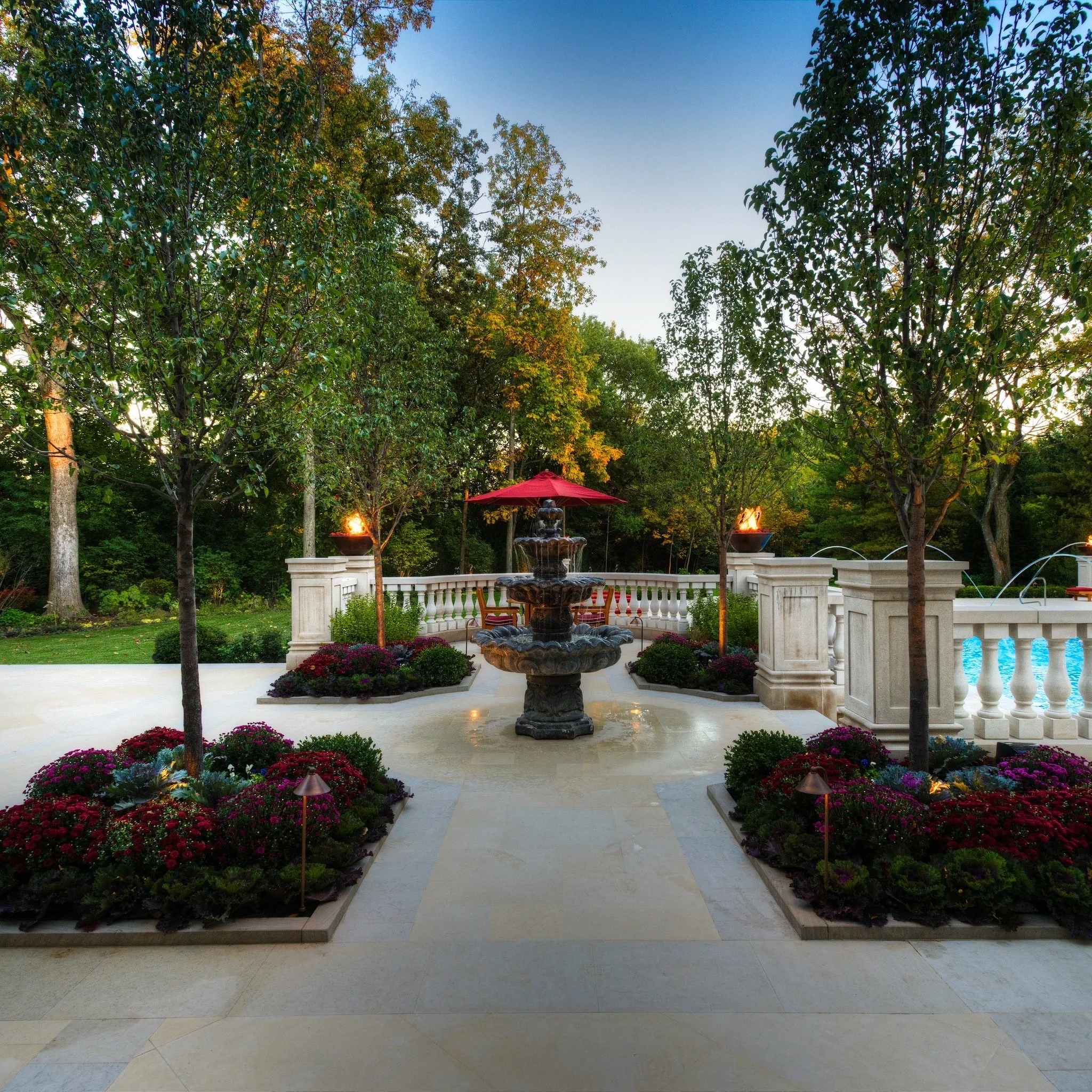 Elegant garden scene with a tiered fountain in the center, surrounded by colorful flower beds and tall trees with green leaves, a fountain with water flowing, and a red patio umbrella in the background.