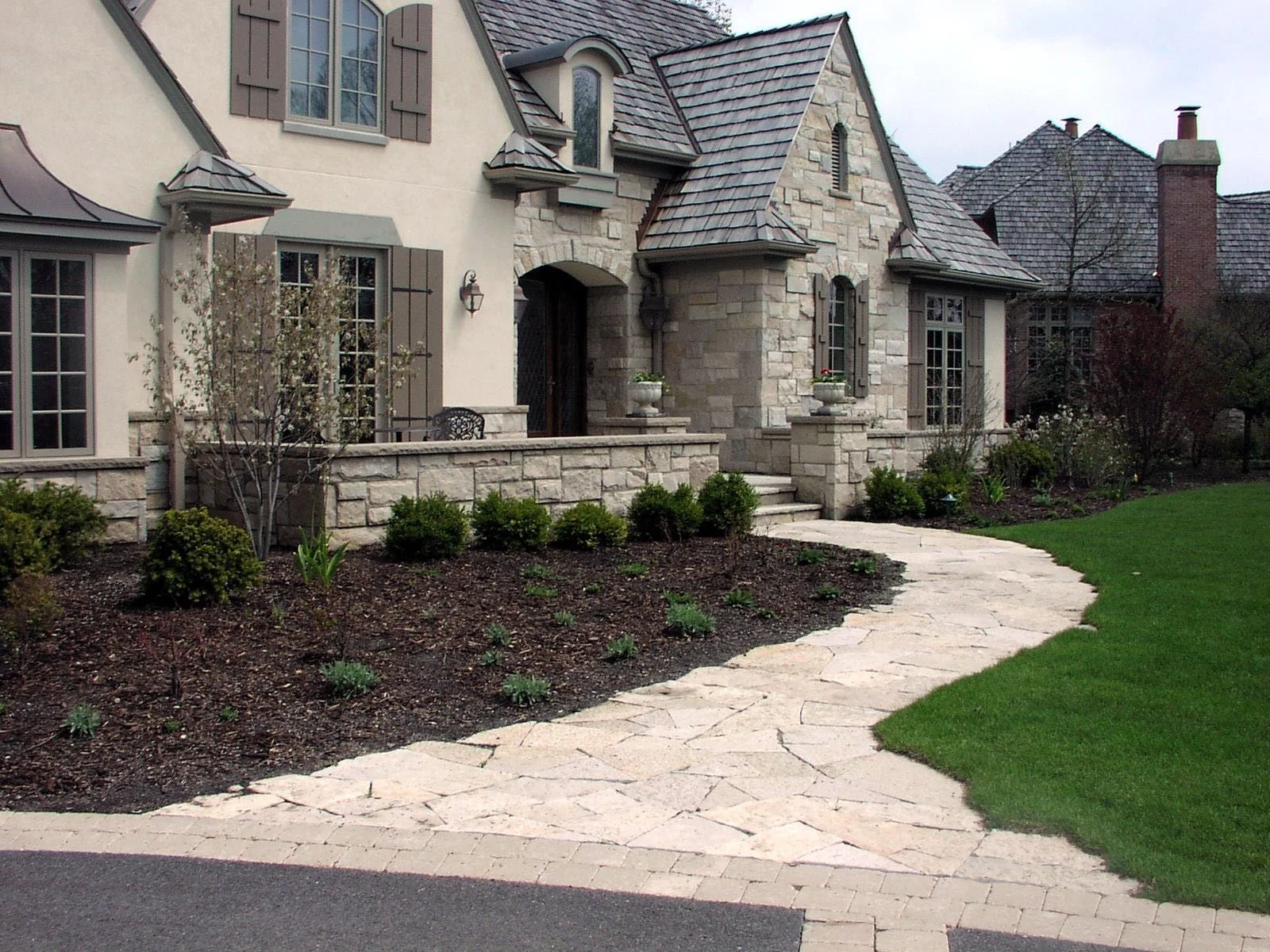 Front view of a large house with a stone and stucco facade, multiple gabled roofs, and a landscaped garden featuring a stone pathway, bushes, and small plants.