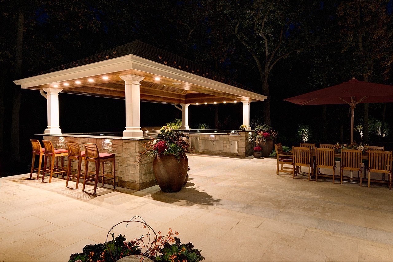 An outdoor patio area at night with a lit-up bar or kitchen structure, surrounded by chairs and tables under a large umbrella, with trees in the background.