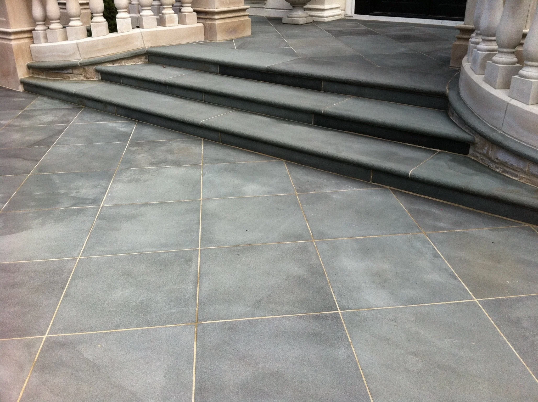 A close-up image of a front porch with stone stairs and a tiled floor. The stairs have three steps, and the floor is made of large, dark gray tiles with light grout lines. The porch has decorative white columns with rounded balusters.