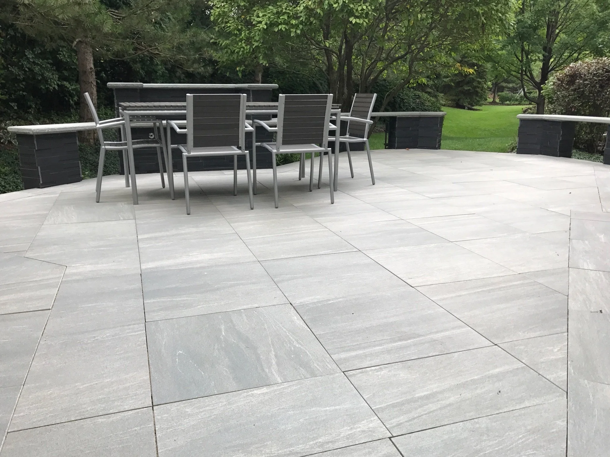 A patio with gray tile flooring, black brick planters, and a set of gray outdoor chairs and a table surrounded by green trees and grass.